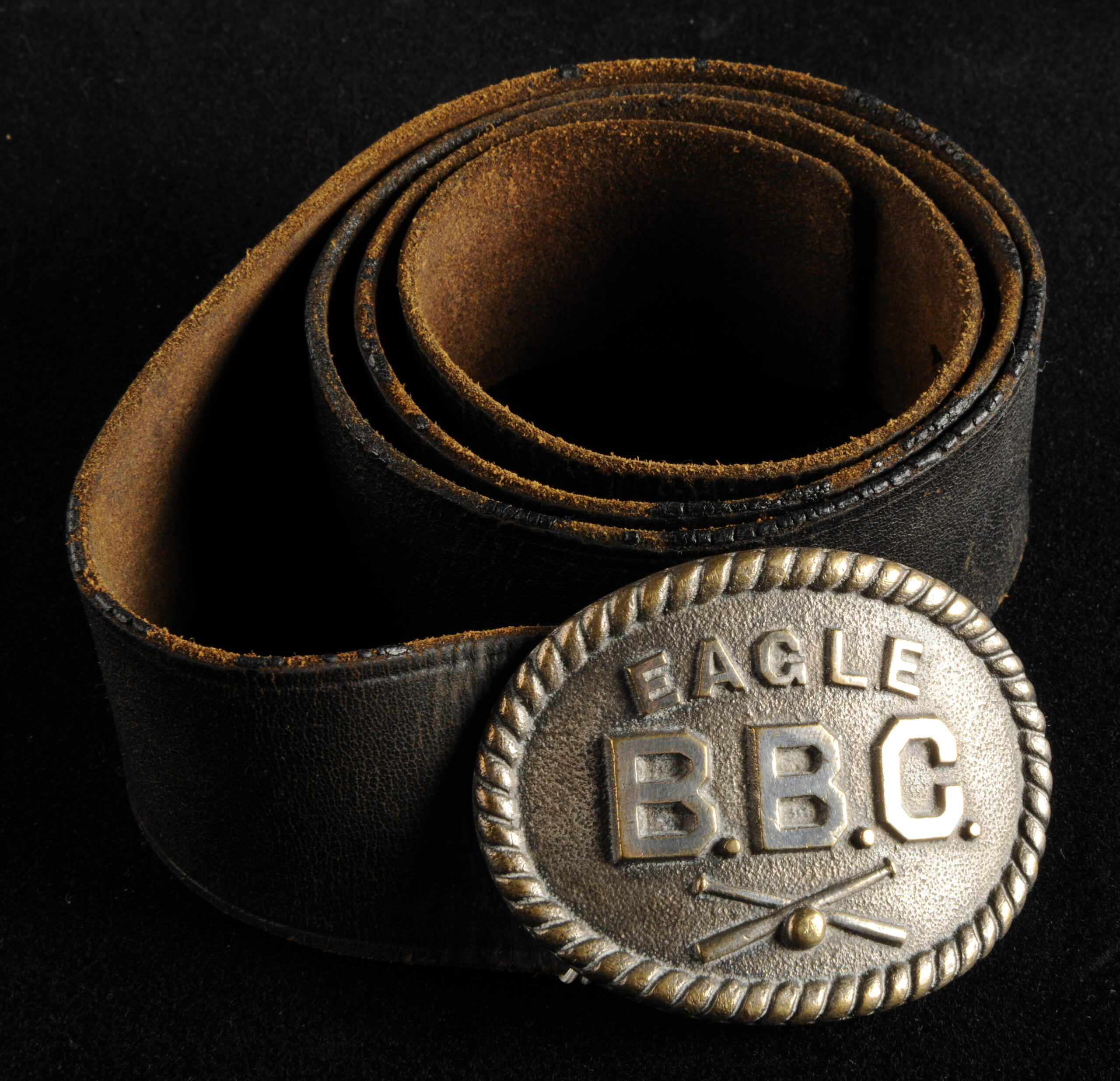 Photograph of a coiled black leather belt and a silver belt buckle against a black background. The belt buckle is oval-shaped with a decorative twisted border. It has text that reads "Eagle B.B.C." Below, there are two baseball bats crossed over each other with a baseball in the center.