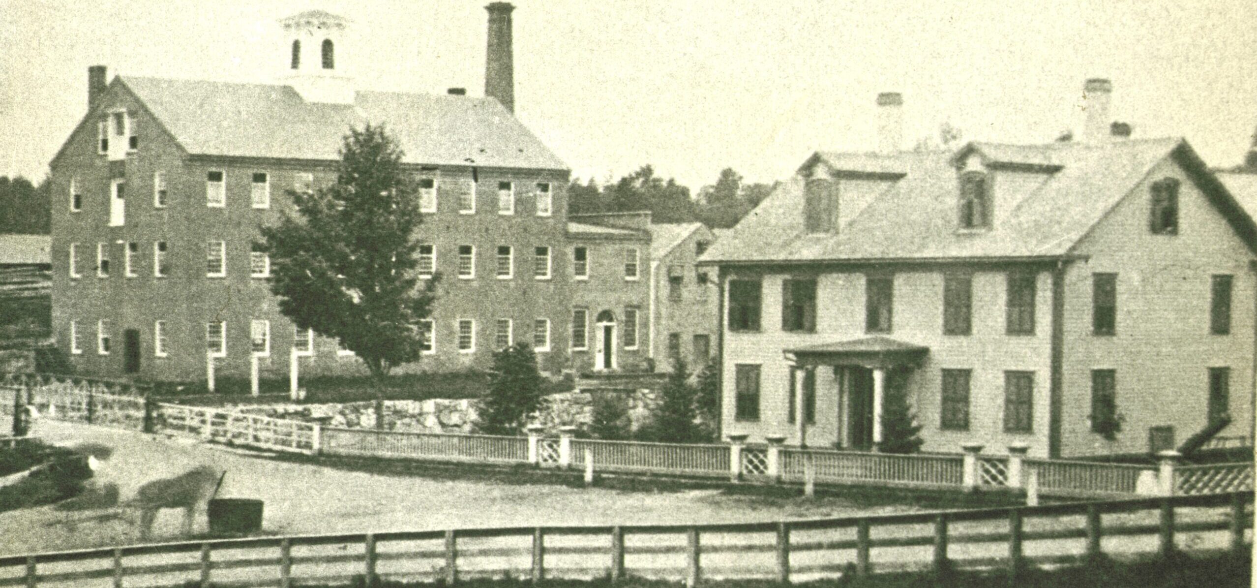 This black-and-white photograph depicts a industrial complex featuring two prominent multi-story buildings situated behind a series of low fences.
On the left stands a large, imposing brick or stone building with four stories, featuring a gabled roof topped with a small white cupola and a tall, dark brick chimney stack rising behind it. Numerous uniform rectangular windows line its facade, and a single tree grows near its front corner. To the right is a lighter-colored, two-and-a-half-story residential-style building with a gabled roof, dormer windows, and a small covered front porch supported by columns.
In the foreground, a dirt or gravel road curves slightly, bordered by a white wooden fence with decorative cross-buck patterns. A horse stands near a watering trough or cart on the lower left side of the path. A lower, rustic wooden rail fence runs horizontally across the immediate foreground, separating the viewer from the scene. Thick trees form a dense background behind the structures