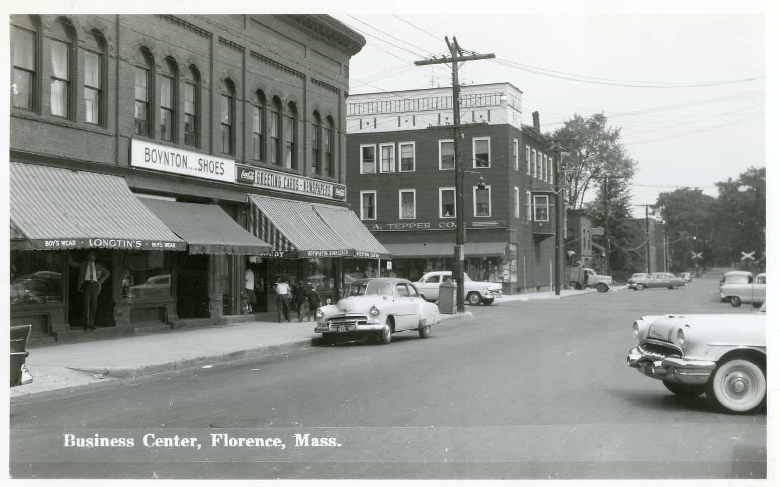 A black and white postcard photograph depicting a street view of the Business Center in Florence, Massachusetts, during the mid-20th century. The perspective is taken from the street, looking down a paved, multi-lane road that bends slightly to the right in the distance, where a railroad crossing with white "X" signs is visible. Vintage mid-century cars are parked along the curbs and situated in the foreground, including a prominent light-colored sedan parked on the left side of the street and the front right fender and whitewall tire of another car in the lower right foreground. On the left side of the street, a multi-story brick commercial building dominates the view, featuring rows of arched windows on its upper floors. The ground floor houses several storefronts protected by large, striped canvas awnings that extend over the sidewalk. Prominent signage above the awnings reads "BOYNTON'S SHOES" and "GREETING CARDS - NEWSPAPERS," while smaller text on the awnings indicates "BOY'S WEAR - LONGTIN'S - MEN'S WEAR." A few pedestrians, including a man standing near a doorway and others walking further down, populate the sidewalk. Across an intersecting side street stands another three-story brick building with the sign "A. TEPPER CO." above its ground-floor storefront. The street is lined with wooden utility poles carrying overhead power lines, and dense trees line the background behind the buildings and along the distant roadway. The bottom left corner of the image features the printed caption text: "Business Center, Florence, Mass."