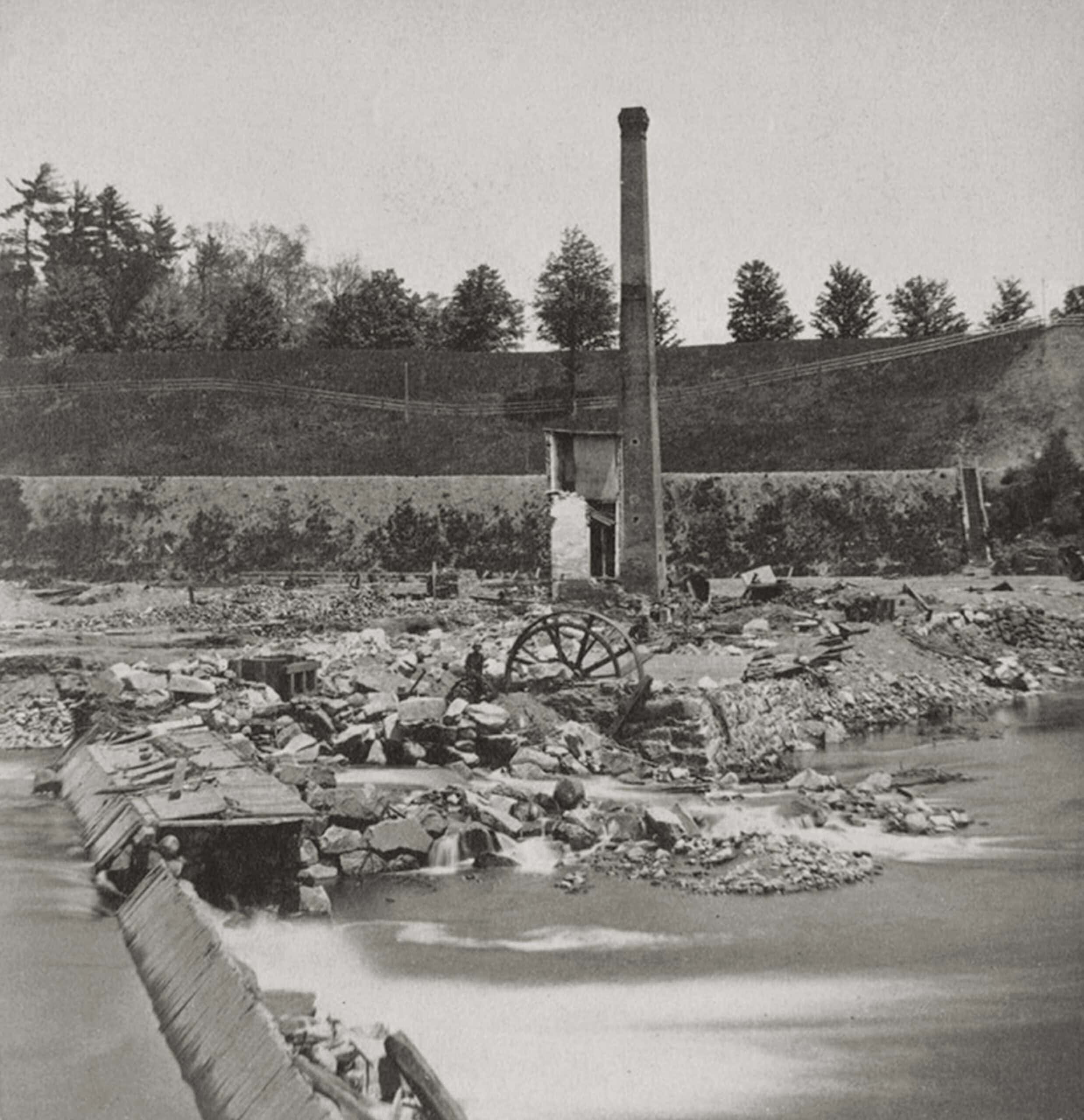A black-and-white photograph of  the remains of a destroyed industrial site. In the foreground, a rushing river flows over a damaged, wooden and stone dam structure, creating blurred white water from a long exposure. The middle ground is filled with piles of rubble, scattered stones, and debris. Rising prominently from the destruction is a tall, slender brick smokestack, standing next to a small, heavily damaged section of a two-story building wall. To the left of the smokestack, a large, broken wooden water wheel sits half-buried amid the rocks. In the background, a steep, grassy hillside or embankment rises up, topped by a row of sparse trees.