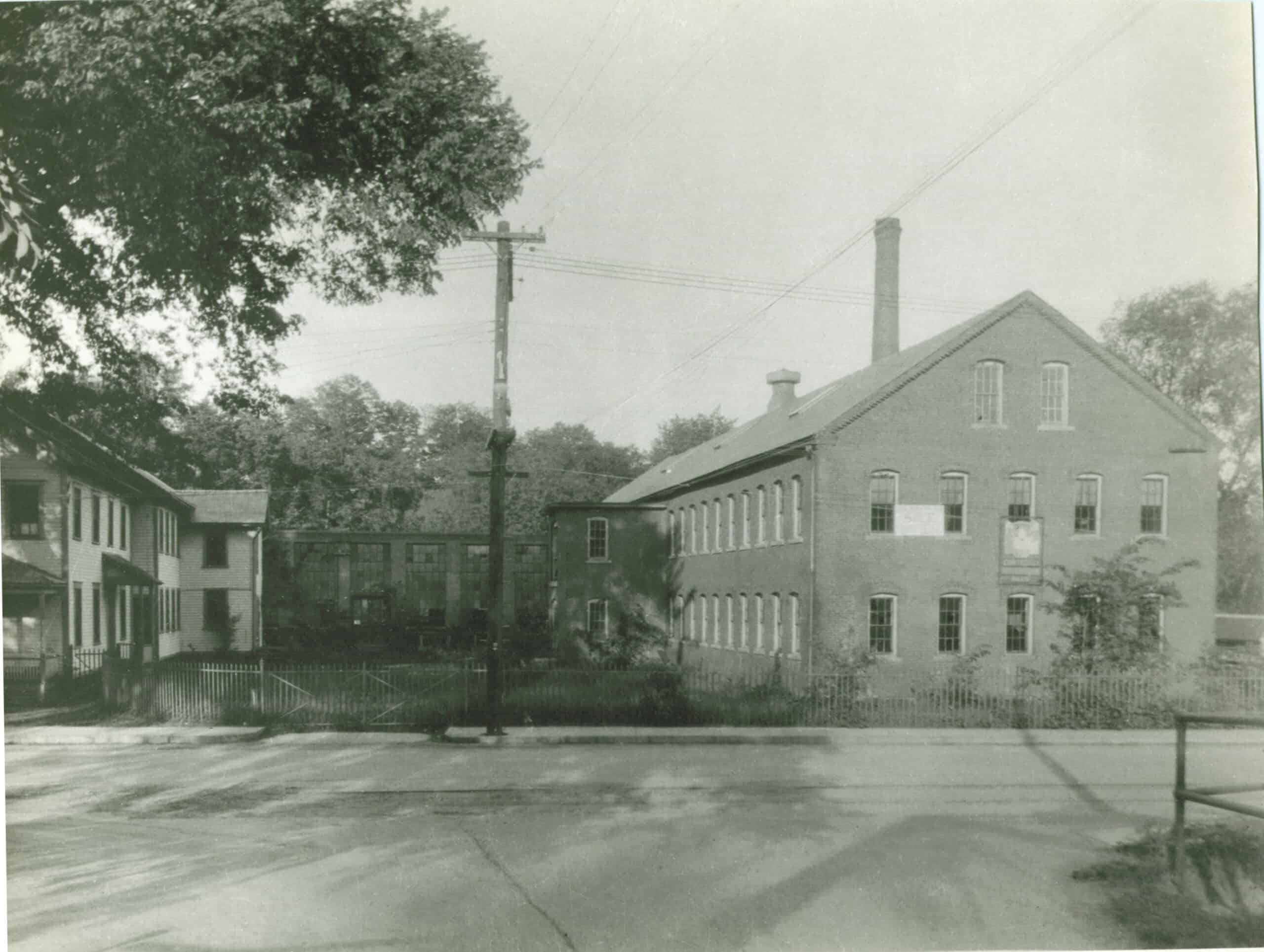A black and white photograph of an industrial complex. On the right, a large two-and-a-half-story brick industrial building dominates the scene, featuring a gabled roofline with a tall brick chimney rising from the rear and a grid of small, multi-pane windows stretching across its facade. A white sign and a dark banner hang on its front exterior walls. To the far left, a separate two-story residential or administrative structure stands with white siding and dark trim around its windows. Connecting these sections in the background is a long, multi-paned glass greenhouse or walkway structure. In the foreground, an unpaved, dusty road runs horizontally, bordered by a low fence and a grassy yard that separates the street from the buildings. A tall wooden utility pole stands near the center of the frame, with power lines stretching across the upper portion of the image. Mature, leafy trees frame the left side and fill the background behind the factory complex.