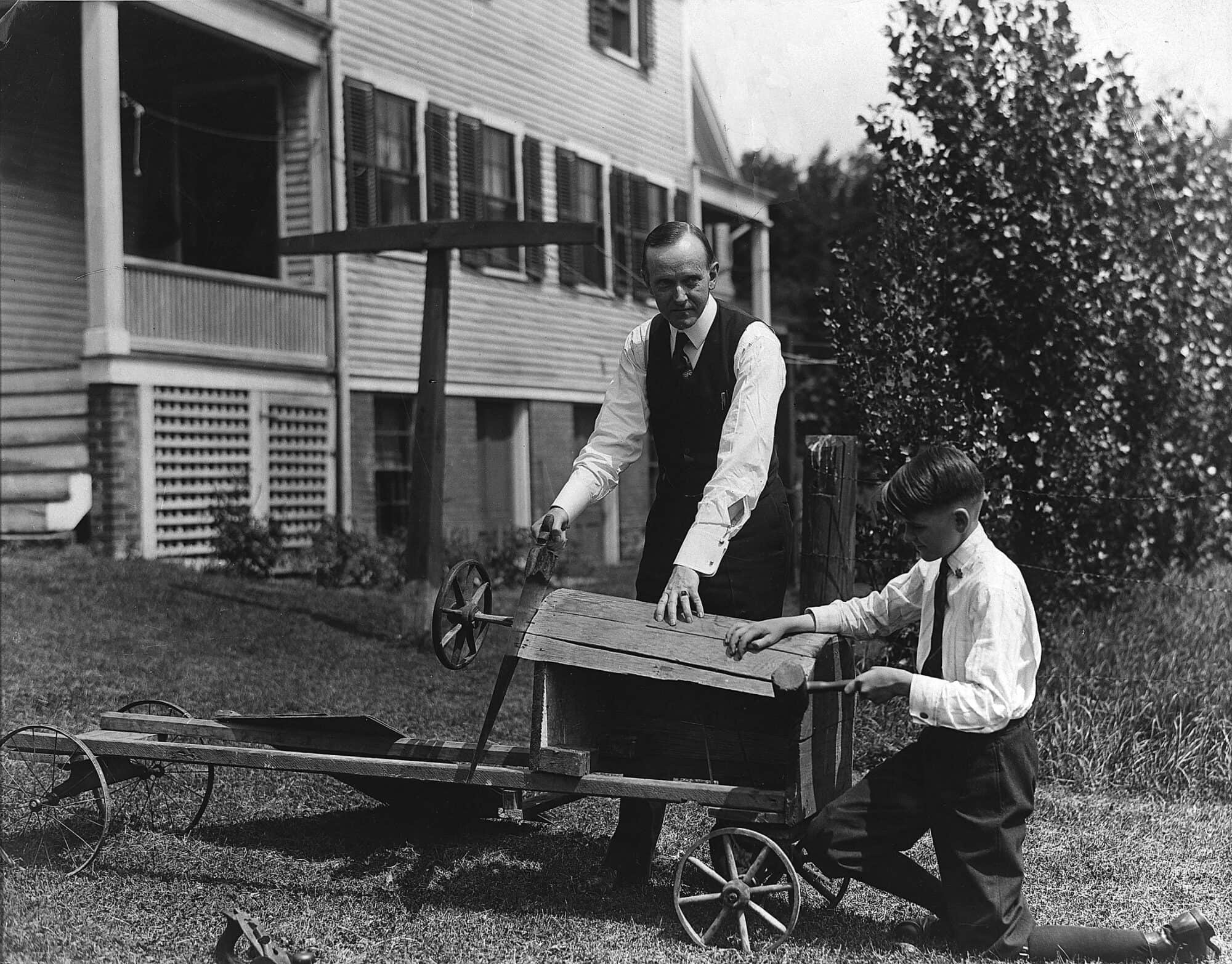 Governor Calvin Coolidge stands behind a soap box racer under construction while holding a saw against the wooden pieces forming the hood. His son Calvin Jr. kneels at the front of the hood with a mallet. Behind them is a view of the backyard and the Massasoit Street house. July 27, 1920.