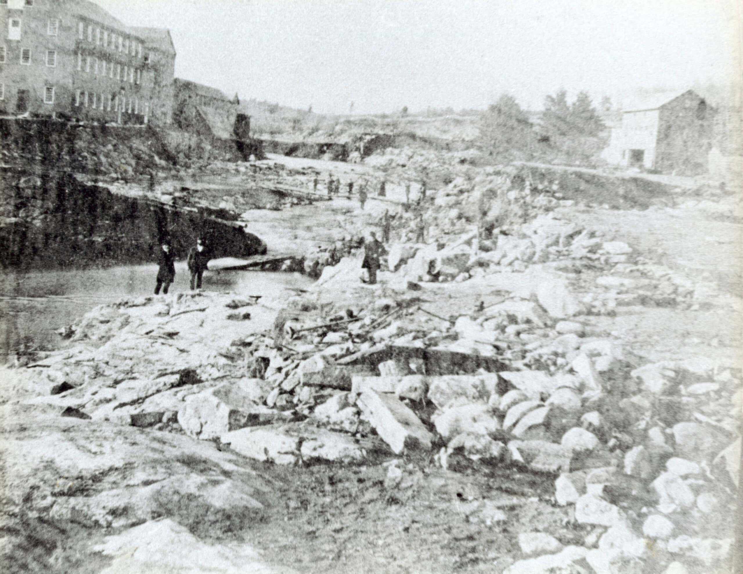 A black and white photograph of a rugged, rocky landscape following a flood. In the foreground and middle ground, the terrain is heavily littered with large broken stones, boulders, and debris. Several individuals in period attire, including dark coats and hats, are scattered across the rocky area, with some standing near the edge of a water channel on the left. In the background on the upper left, a large multi-story brick or stone industrial building sits atop an elevated bank. Across the riverbed toward the right, a smaller stone structure is visible amidst trees and uneven ground.
