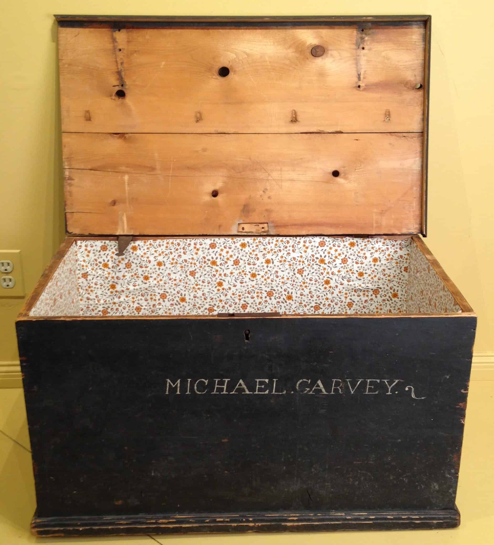 Photograph of a wooden chest with its lid open. The exterior of the chest is painted black with the name "Michael Garvey" written on it. The interior of the lid is unpainted wood. Inside the chest is painted or wrapped in a orange and white floral print.