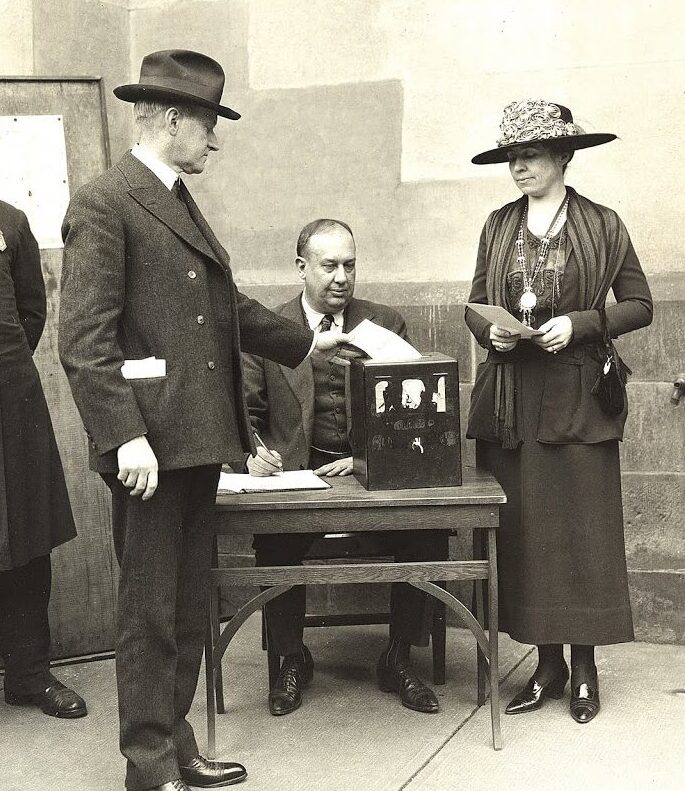 Black-and-white photograph of Calvin and Grace Coolidge standing outside at the ballot box. A man sits infront of a table that the box is on observing as Calvin Coolidge places his ballot in.