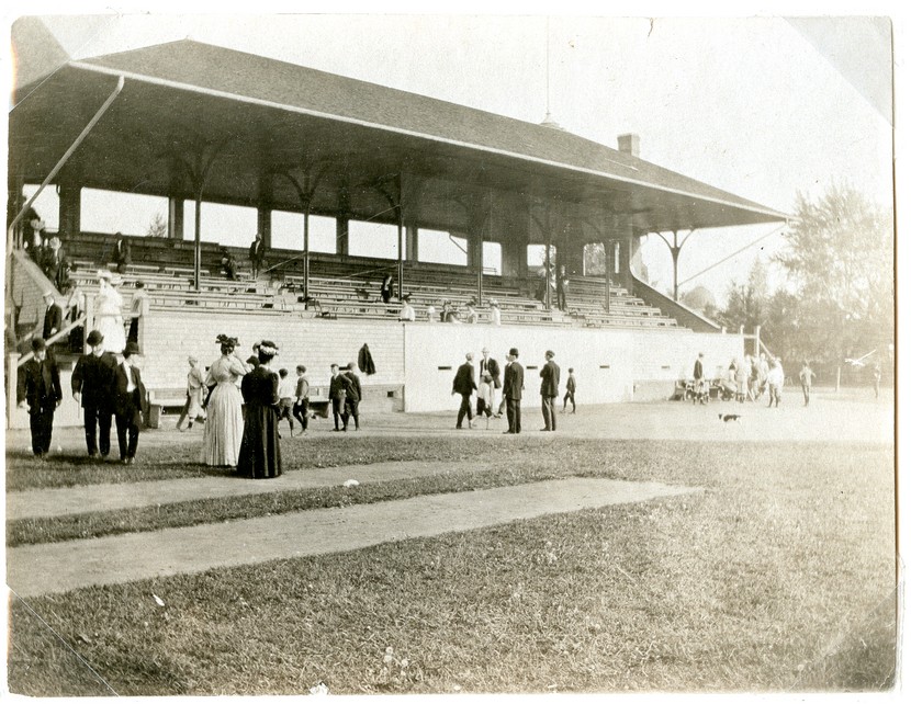 Black and white photograph of a grandstand with a brick wall, elevated bleachers, and a roof held up by metal pillars. There are a few people in the stands. In the foreground, there is a field, some of it grassy and some of it dirt, with several people walking around it in 1890s clothing. 