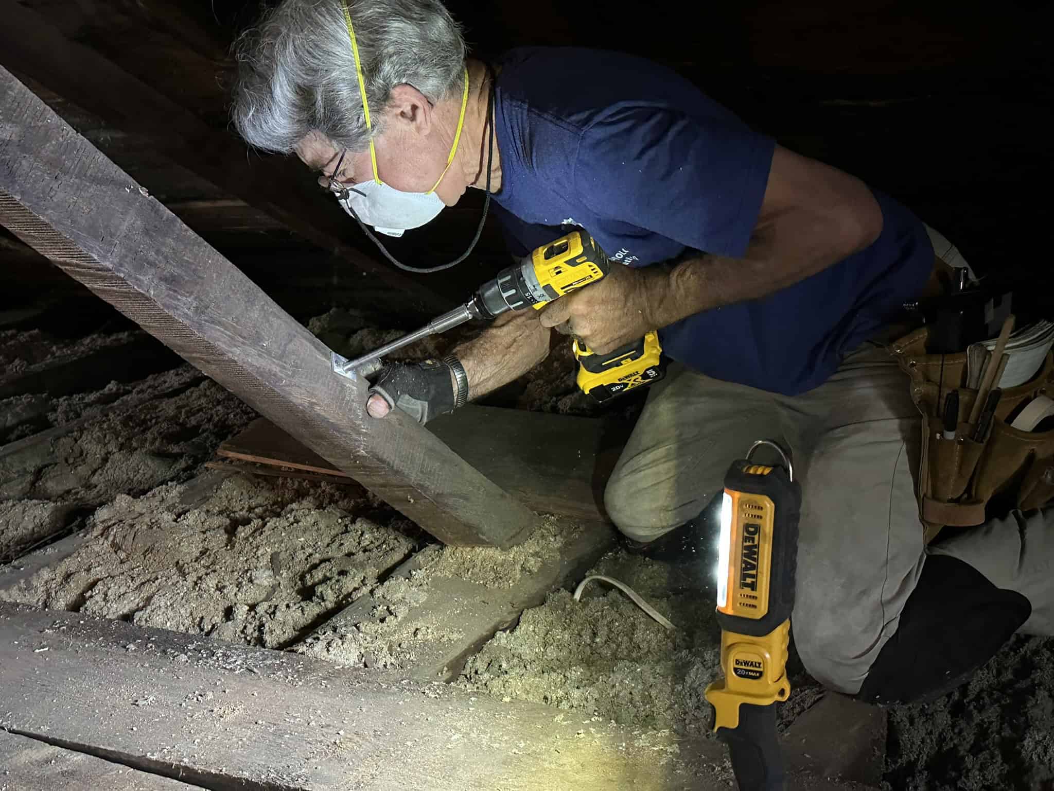 Image of a man working in a dimly lit, narrow attic space filled with loose-fill insulation, using a yellow and black cordless drill on a structural wooden beam. The man, who has gray hair, glasses, a face mask with yellow straps, and a dark blue t-shirt, is positioned on the right side of the frame, facing left. He is kneeling on the insulation-covered attic floor, wearing gray work pants and a leather tool belt equipped with multiple pockets on his left hip. With gloved hands, he holds the DeWalt cordless drill, which is angled downward as he drives a screw into a metal plate attached to a thick, weathered diagonal wooden beam. In the lower right foreground, a yellow and black DeWalt LED work light stands upright on the floor. The surrounding attic space is dark, with exposed wooden rafters visible in the background and thick layers of gray insulation covering the floor joists.