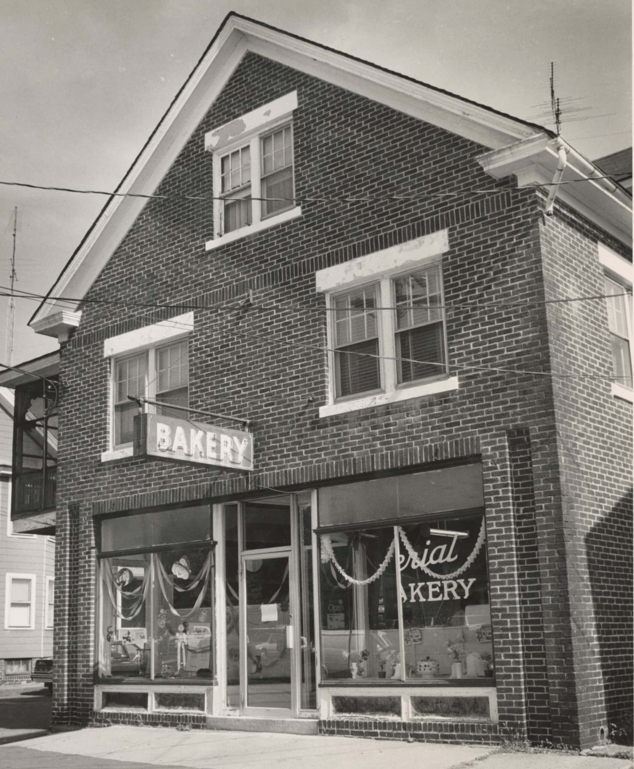 Black-and-white photograph of Imperial bakery. The building features a red brick facade with a prominent triangular gabled roofline at the top, trimmed with white molding. Multi-paned double windows with thick white lintels are visible on the second and third floors. On the ground floor, a large storefront glass window on the right features cursive script lettering reading "...arial BAKERY" decorated with a string of white garland. A matching large window on the left displays decorated cake stands and bakery items through the glass. A central glass entry door stands slightly recessed, flanked by the two windows. Projecting from the left side of the brick wall above the windows is a rectangular business sign that reads "BAKERY" in bold block lettering.