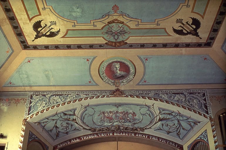 Photograph of a Renaissance-style fresco with pale blues, seafoam greens, muted tans, and reddish-orange accents. In the center of the ceiling is a classical Romanesque bust of a figure set infront of an reddish-orange background framed by a wreath.  In a section that appears to be draped over a door, it reads: "ABOVE ALL THINGS, TRUTH BEARETH AWAY THE VICTORY."