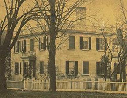 A black-and-white photograph of the Damon House on Bridge Street. The house is a white squared building with black shutters. An ornate entrance way can be seen in view. Two bare trees stand infront.