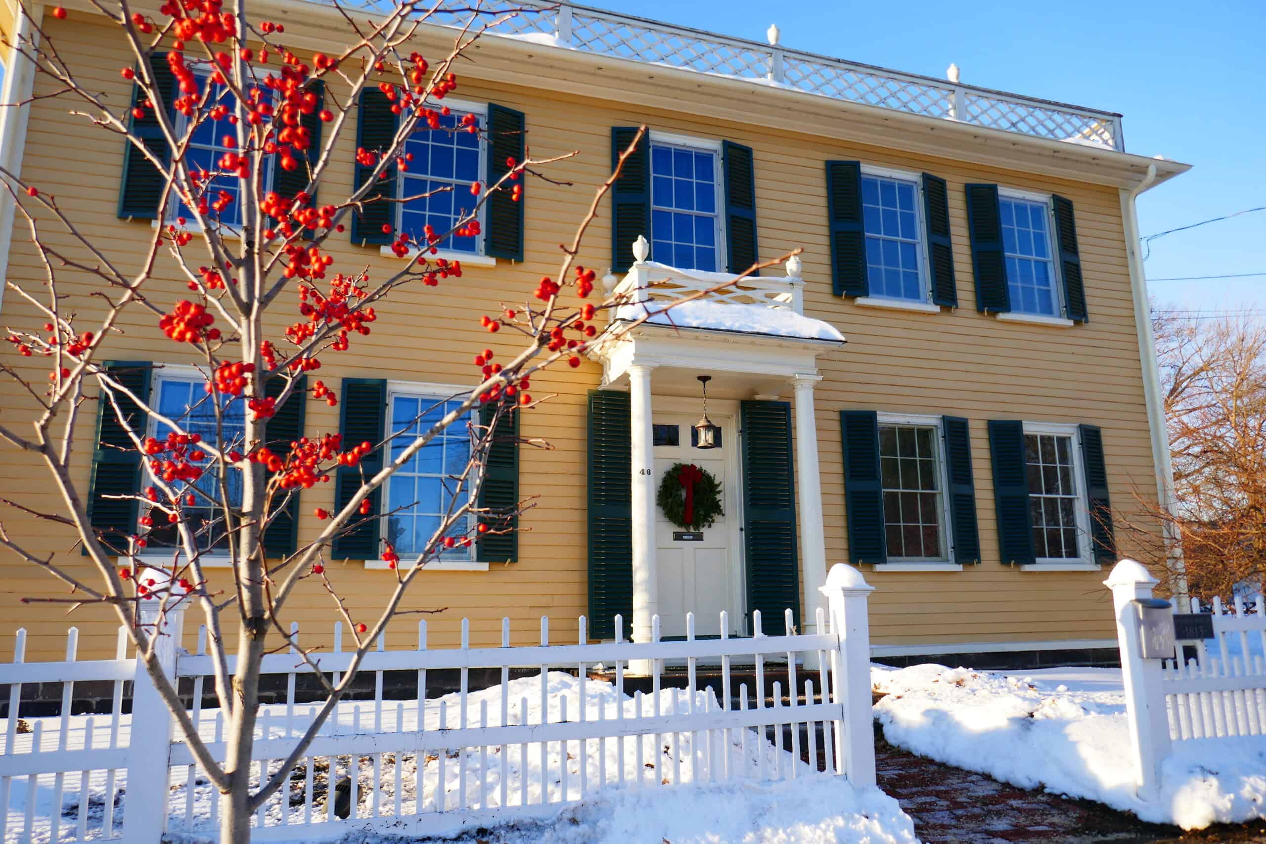 A large, two-story historic house with light yellow horizontal siding features dark green shutters on its numerous multi-pane windows. The central front entrance is framed by a white-columned portico covered with a dusting of snow, and a festive green wreath hangs on the white front door. A white picket fence runs along the snow-covered front yard, partially obscured on the left by a slender, bare tree branches laden with bright red berries. The roofline is accented by an decorative white rooftop railing against a clear blue sky, while patches of snow cover the ground and the top of a mailbox post to the right.