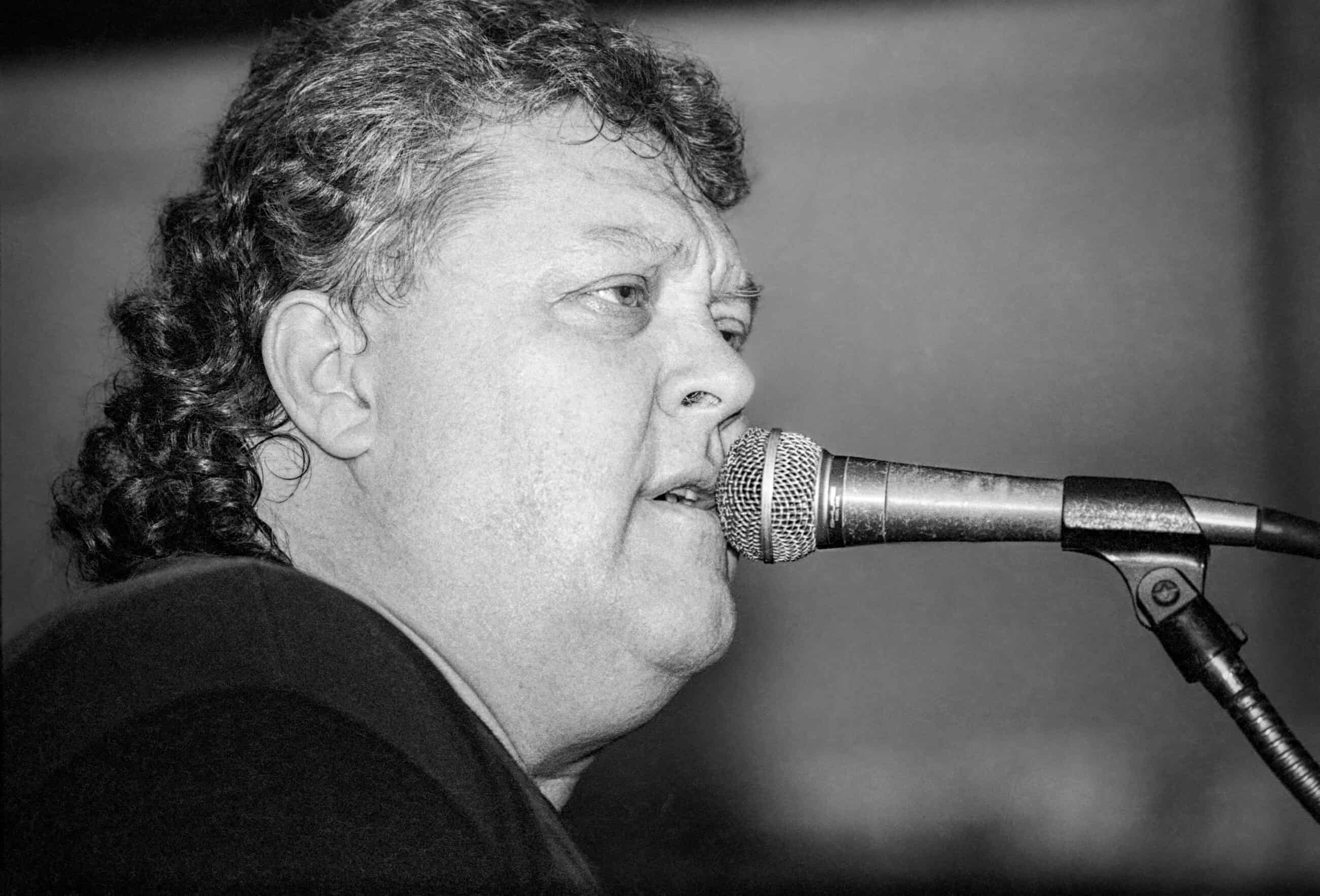 Black-and-white photograph of a middle-aged man with curly dark but graying hair speaking or singing into a microphone.