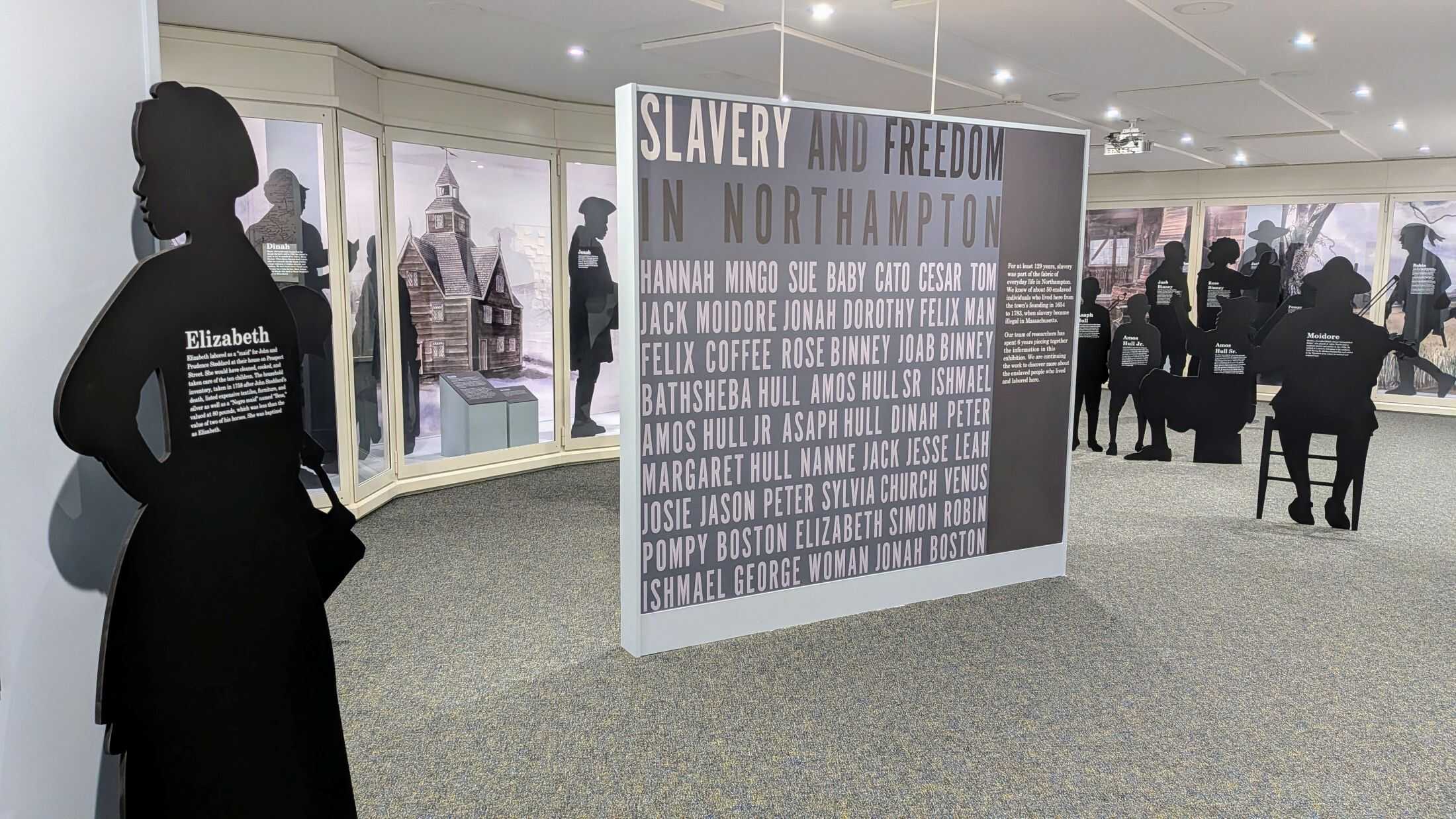 Photograph of a well-lit, small exhibit space with a gray rug and white and cream walls. Exhibit cases are built into the walls and show different scenes of enslaved people laboring in 18th century Northampton Scattered around the room are cardboard cutouts of silhouettes of people, the silhouettes are black and have white text on them, saying the name of the person it is representing and some information about them (None are readable in this image). In the center of the exhibit space is a tall and thin board, only a few feet below the ceiling. The board reads: "SLAVERY AND FREEDOM IN NORTHAMPTON" with the names of some of the enslaved people who lived in Northampton, along with some text that is unreadable in the image.