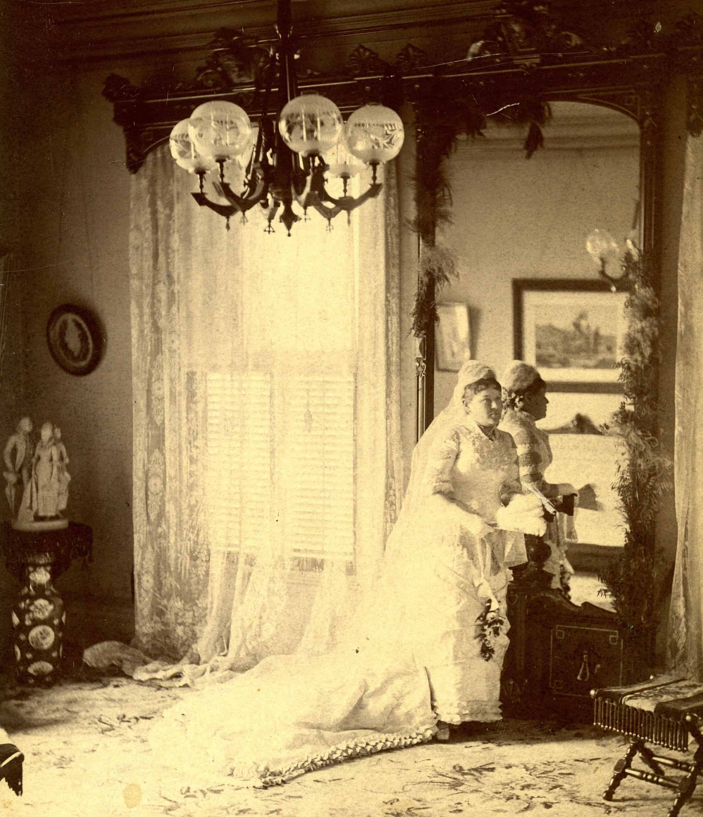Black-and-white photograph of a woman standing infront of a mirror in her wedding dress and veil. Her dress has a long train.