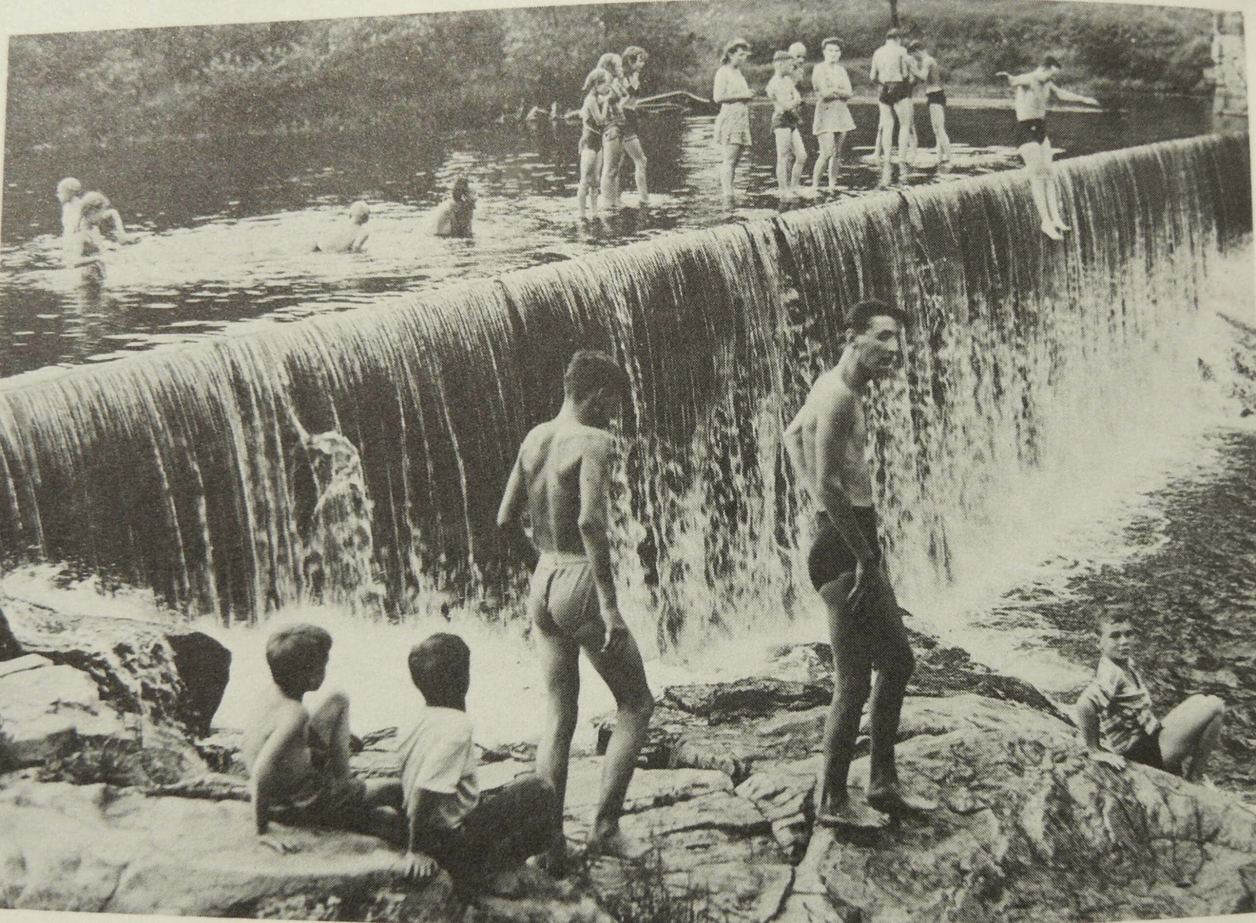 Black-and-white photograph captures a large group of children and teenagers swimming and playing around a wide river dam on a summer day. The concrete dam cuts diagonally across the center of the frame, with a steady, white curtain of water cascading down its face into a rocky pool below. Along the very top edge of the dam, a line of about a dozen kids in mid-20th-century swimwear stand and wade in the shallow water, while on the far right, a boy balances on the lip, preparing to jump or dive into the deep water below. In the calm water behind the dam, several more children can be seen swimming and wading. The foreground features a rugged, rocky riverbed below the falls where two young boys sit together on a boulder facing away from the camera, watching two older, shirtless boys stand nearby near the splashing water. On the far lower right, a young boy wearing a plaid shirt sits alone on a flat rock, looking directly at the camera, while a dense, out-of-focus forest line frames the upper background.