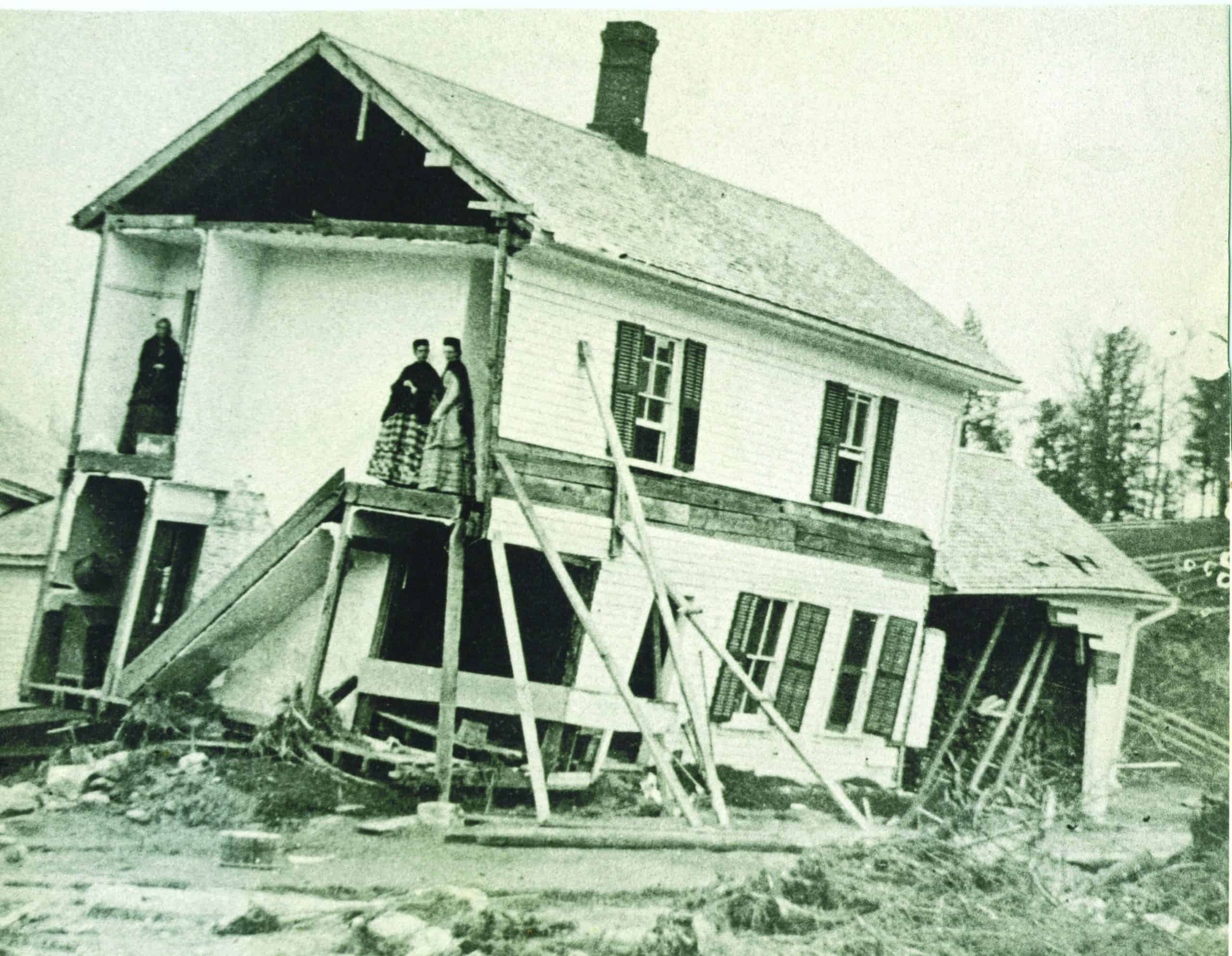 A black-and-white photograph of a two-story wooden house heavily tilted to the right after severe structural damage. The left section of the house has been sheared away, entirely exposing its interior layout. On the second floor of this exposed section, two women dressed in long, dark, period-style dresses with patterned skirts stand near the edge of the floorboards, looking out. To their left, another individual in a dark coat stands in the doorway of a separate, higher room. The ground floor beneath them is completely destroyed, leaving the upper level precariously supported by a chaotic arrangement of thick wooden beams and makeshift stilts angled into the muddy, debris-strewn ground. The remaining intact portion of the white house features multi-paned windows with dark shutters, a shingled roof, and a dark brick chimney rising from the center. Debris and mud cover the foreground.