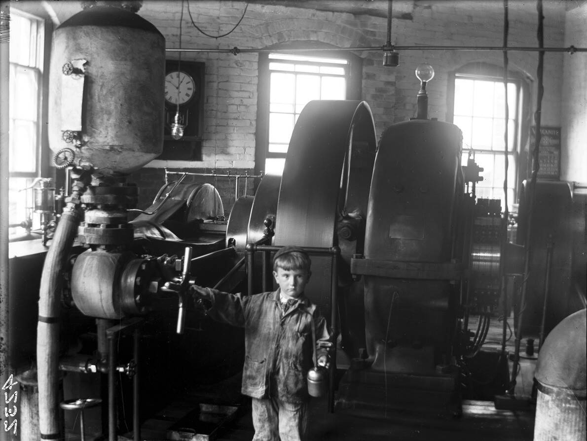 A black-and-white photograph of a toddler or very young boy working in the Norwood Engineering building. He is looking toward the camera man while holding on to large machinery.