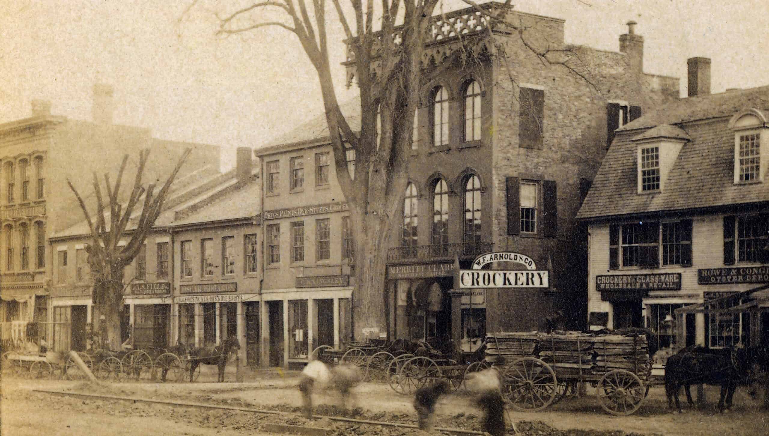 Black-and-white photograph captures a 19th-century American street scene with horse-drawn wagons parked in front of rowed brick commercial buildings. : A block of connected federal and Victorian-style brick storefronts stands prominently in the center. A distinct three-story building features a large storefront sign reading "W.F. ARNOLD & CO. CROCKERY" on the ground level. The upper floors display elegant arched multi-pane windows and an ornate Gothic or Italianate-style cornice line along the roof. To the right, a smaller building with a dormer window bears a sign reading "CROCKERY, GLASS WARE" and "HOWE & CONANT OYSTER DEPOT". To the left, several multi-story merchant buildings line the street.