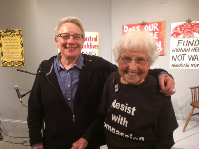 Photo of two women with arms around each other smiling at the camera, in a gallery with political posters hung on the wall behind them
