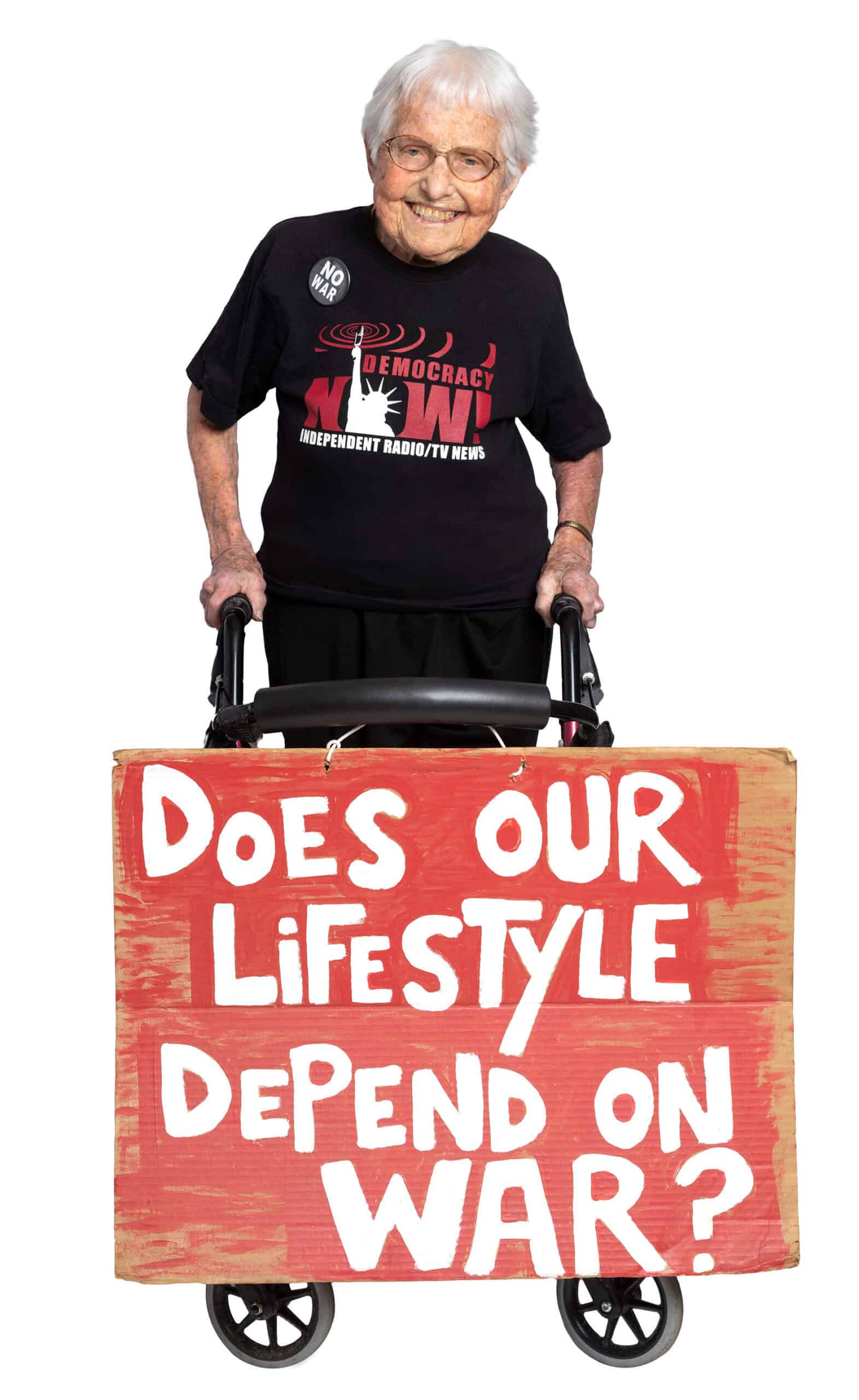 Photograph of an elderly woman standing with a walker on which hangs a protest sign reading "Does our Lifestyle Depend on War?' She is wearing a t-shirt reading 'Democracy Now! Independent Radio/TV News' and a button reading 'No War'