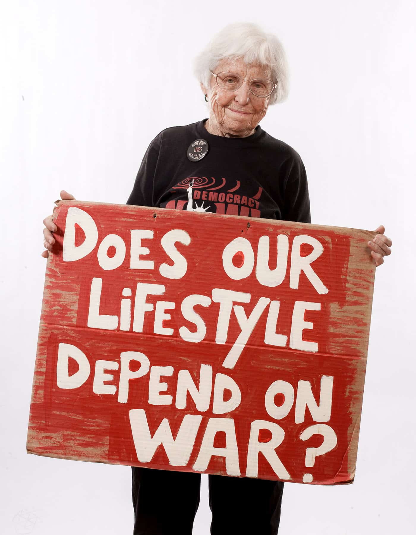 Photograph of an elderly woman holding a protest sign reading 'Does Our Lifestyle Depend on War?'
