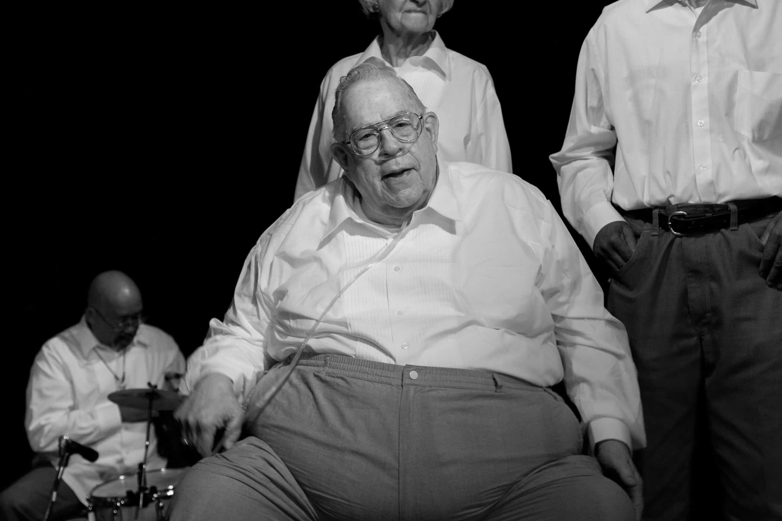 Black-and-white photograph of a matured man sitting and looking past the camera. He has a breathing tube in his nose and is wearing a white buttoned down shirt and dark dress pants. His white hair is swept away from his face.