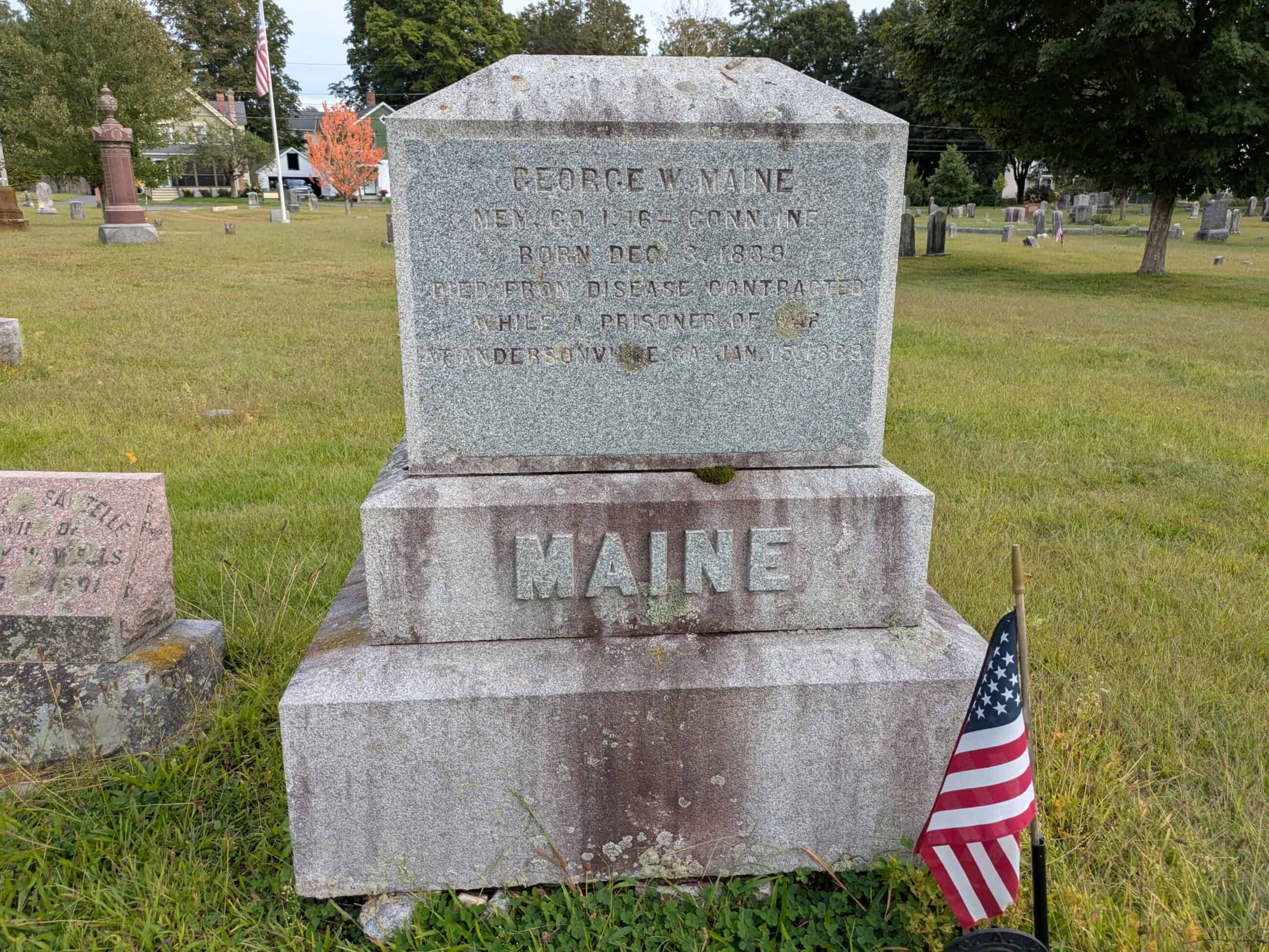 Photograph of the Maine gravestone with a small American flag stuck into the ground next to it, it reads:  "GEORGE W. MAINE / MEM CO. I. 16 - CONN. INE. / BORN DEC 3, 1839 / DIED FROM DISEASE CONTRACTED / WHILE A PRISONER OF WAR... [Rest not intelligible]."