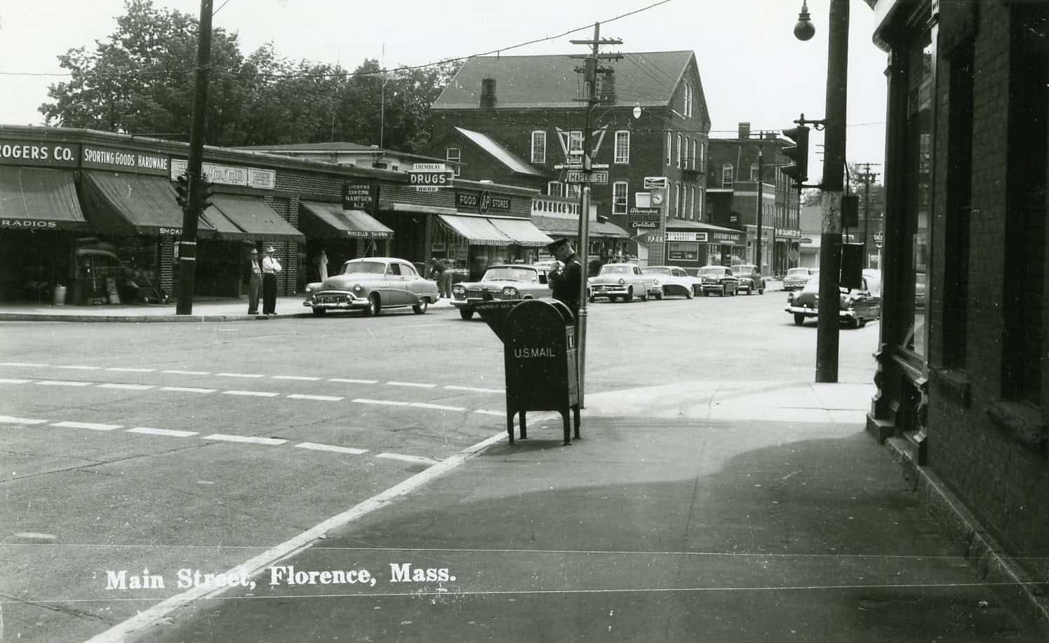 Black-and-white photograph of Goodwin Block at the intersection of Maple Street and Main Street. Storefronts are in view, and cars can be seen parked on the sides of the roads. From left to right, the visible signs on the street are: "F.J. Rogers Co. Sporting Goods Hardware", "Radios". "Smoking", "MurDuffs","Liquors Tavern Hampden Ale", "Purcells Tavern", "Tremblay Drugs", "U.S. Mail", "A&P Food Stores", "Miss Florence Diner", "Maple Street", "Budweiser", "Alexander's ... Cocktails", "Park"