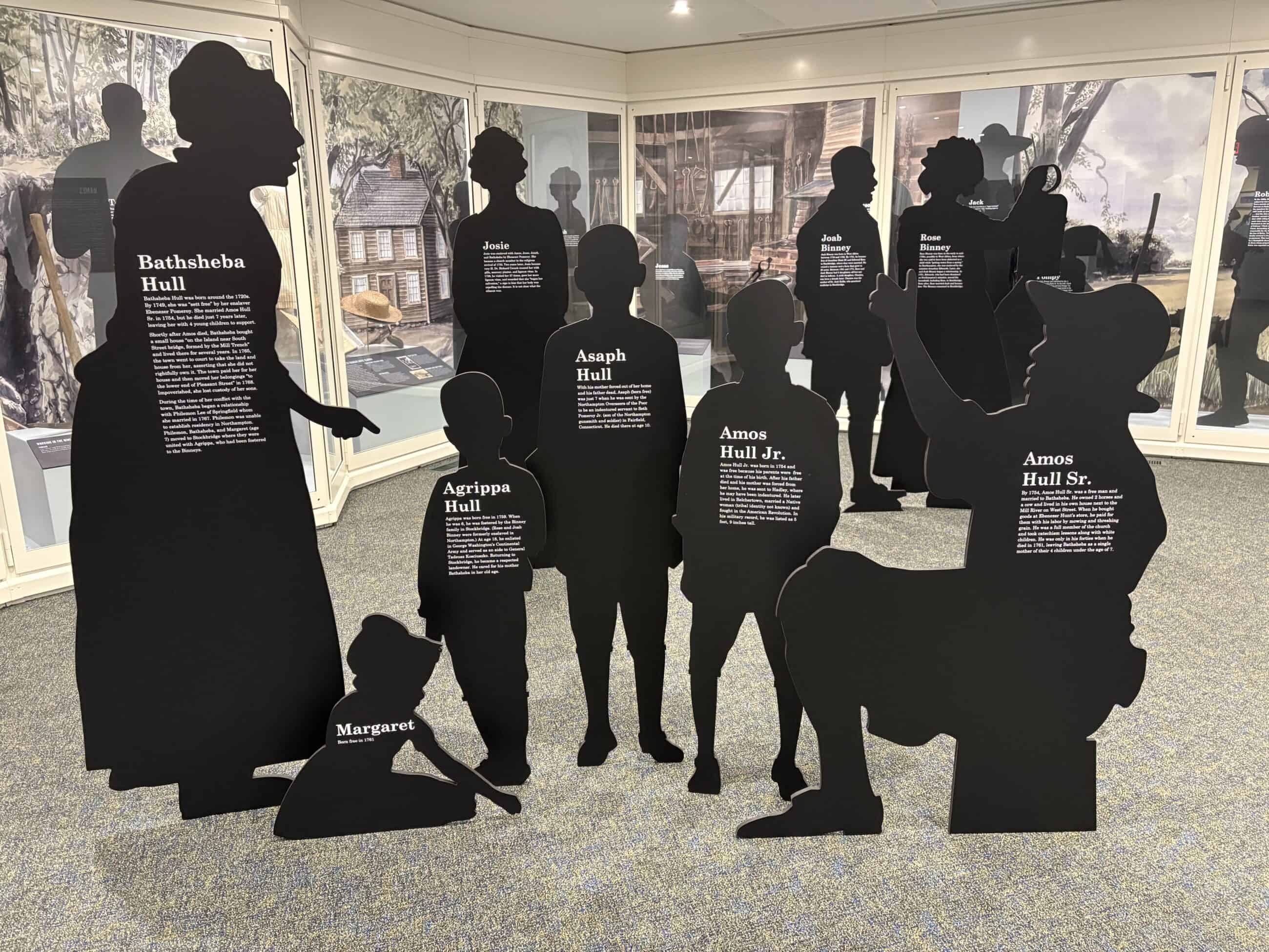 Photograph of all of the cardboard cut out silhouettes of the Hull family created for the Slavery in Northampton exhibit at Historic Northampton. Each exhibit has a name and text for the person they are supposed to represent. The exhibit cases can be seen lining the wall in the background.