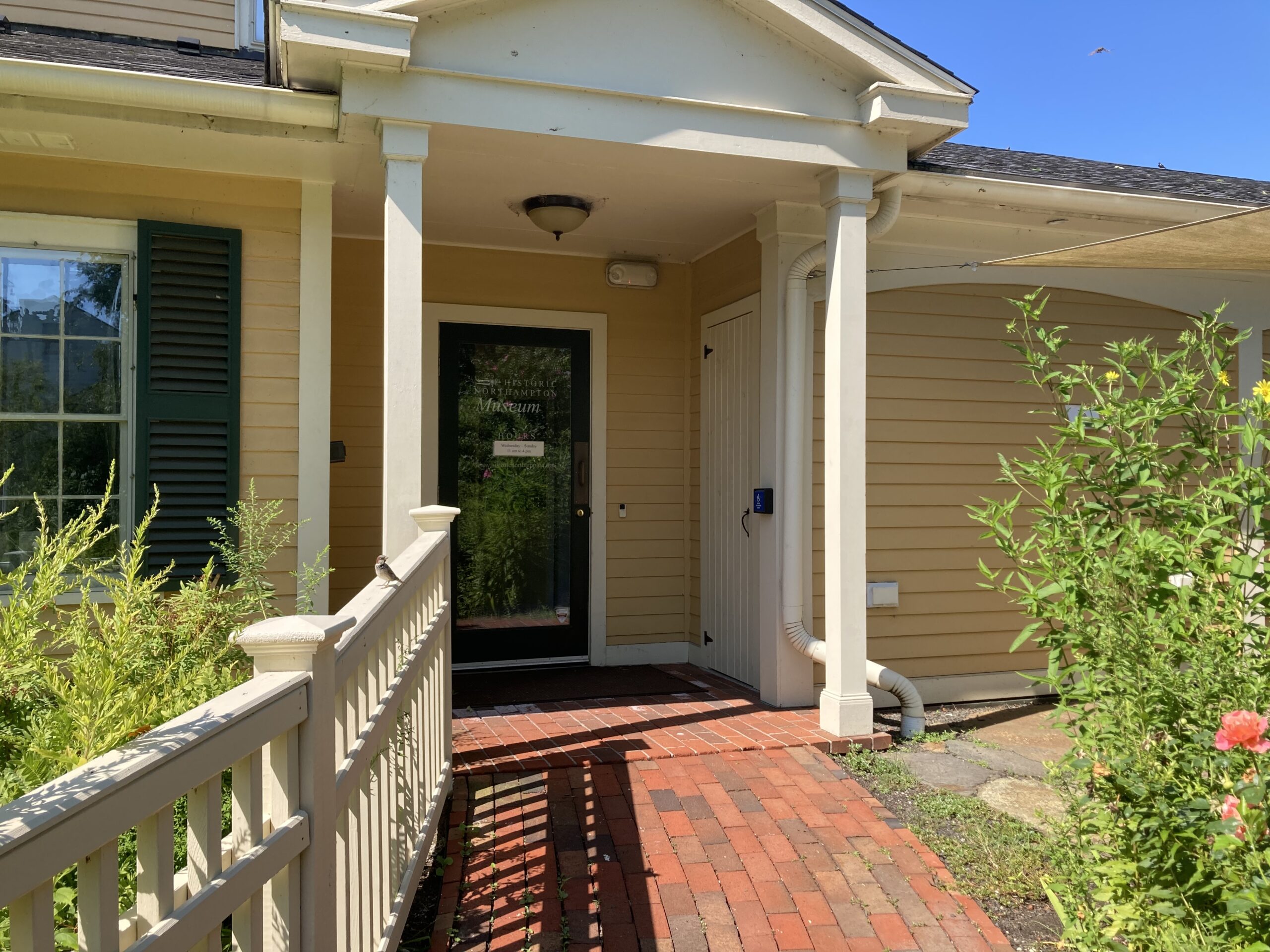 Photograph of a yellow building with dark green shutters, cream-colored trim, and a glass door under a portico with a triangular pediment. There is a red brick sloped pathway leading to the door and cream-colored fence to the left side of the railing, with a small sparrow perched on the fence. There are bushes with pink and yellow flowers on either side of the pathway.
