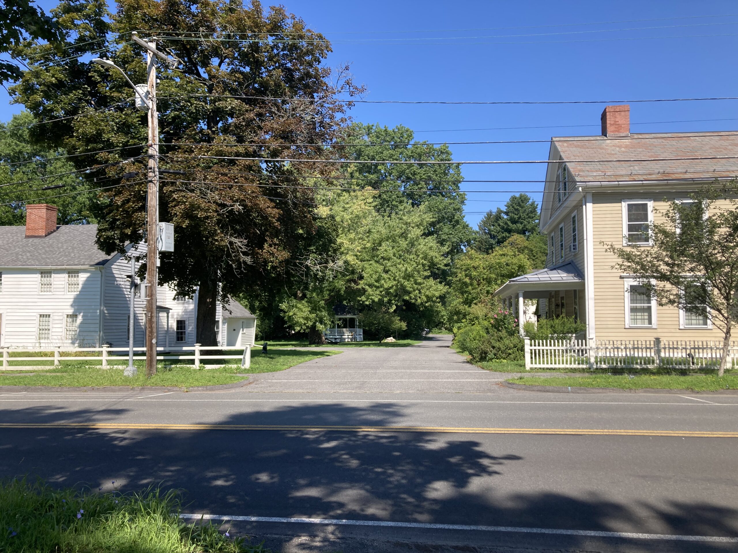Photograph showing a the street, driveway entrance, and driveway between two historic buildings, one white and one beige. In the background is a white gazebo and several trees.