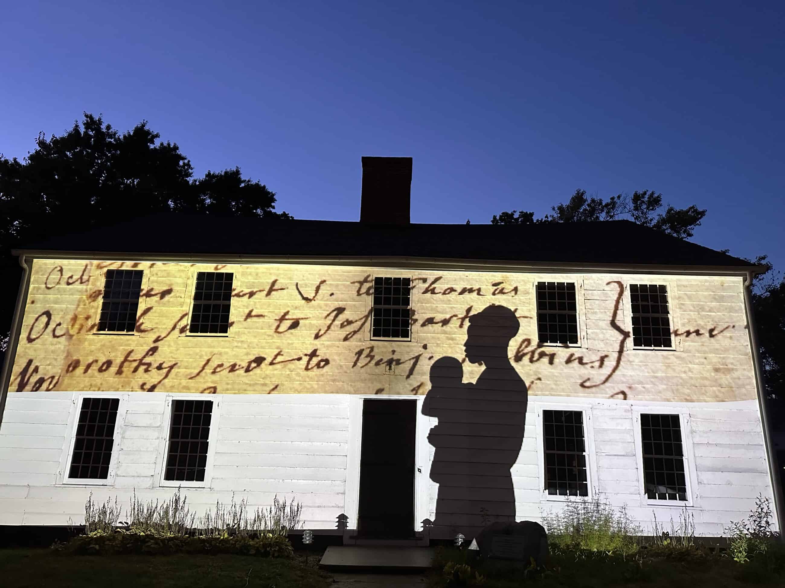 Photograph of the Parsons House owned by Historic Northampton with the image of a silhouette of an enslaved woman holding a baby with a yellowed document in the background projected on it.