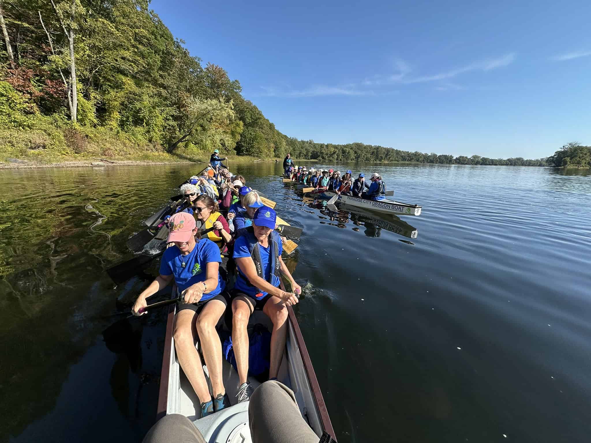 Photo of Connecticut river with two long boats pull of people paddling. There is a tree-filled shoreline and blue sky in the background.
