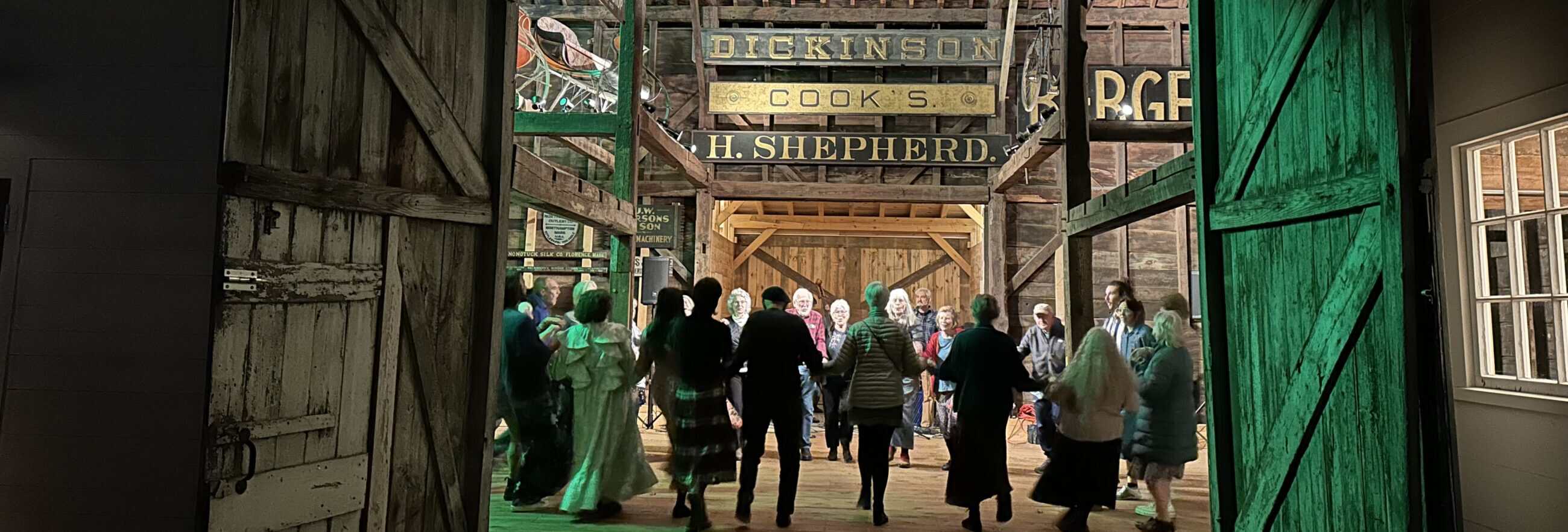 Photograph of group of people dancing hand-in-hand in a circle inside a rustic wooden barn, framed by large, open barn doors. The heavy, weathered wooden doors frame the shot on the left and right. The door on the right is illuminated with a vibrant green light. Through the doorway, a group of about fifteen people of various ages are holding hands and dancing in a ring on a light-colored wooden floor. Most have their backs to the camera. They wear casual clothing, including sweaters, skirts, and jackets. The barn interior features exposed timber framing and a high ceiling. Several vintage wooden signs are mounted high on the back wall, including ones that read "DICKINSON," "COOK'S," and "H. SHEPHERD."