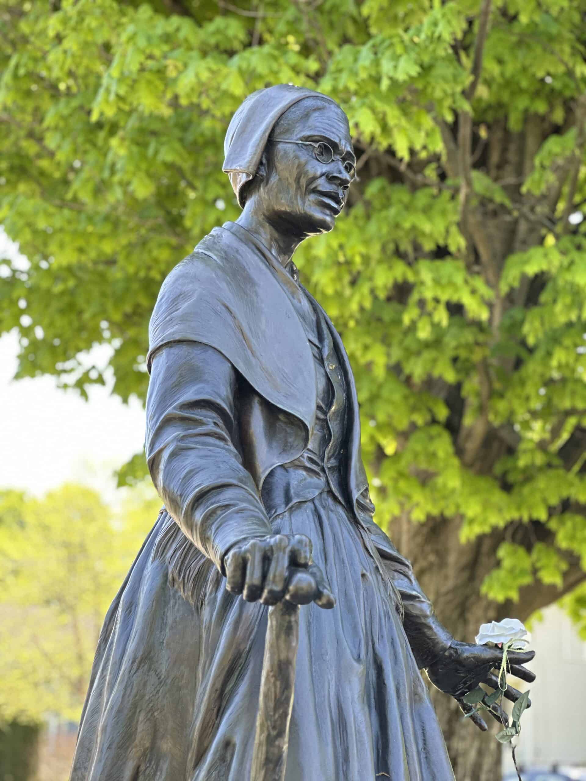 Photograph of a side-view of the Sojourner Truth statue in Florence, Mass. She is wearing a dress with a jacket draped over her shoulders. She has on a bonnet. In one hand she is holding onto a cane and in the other someone has placed a real white flower.