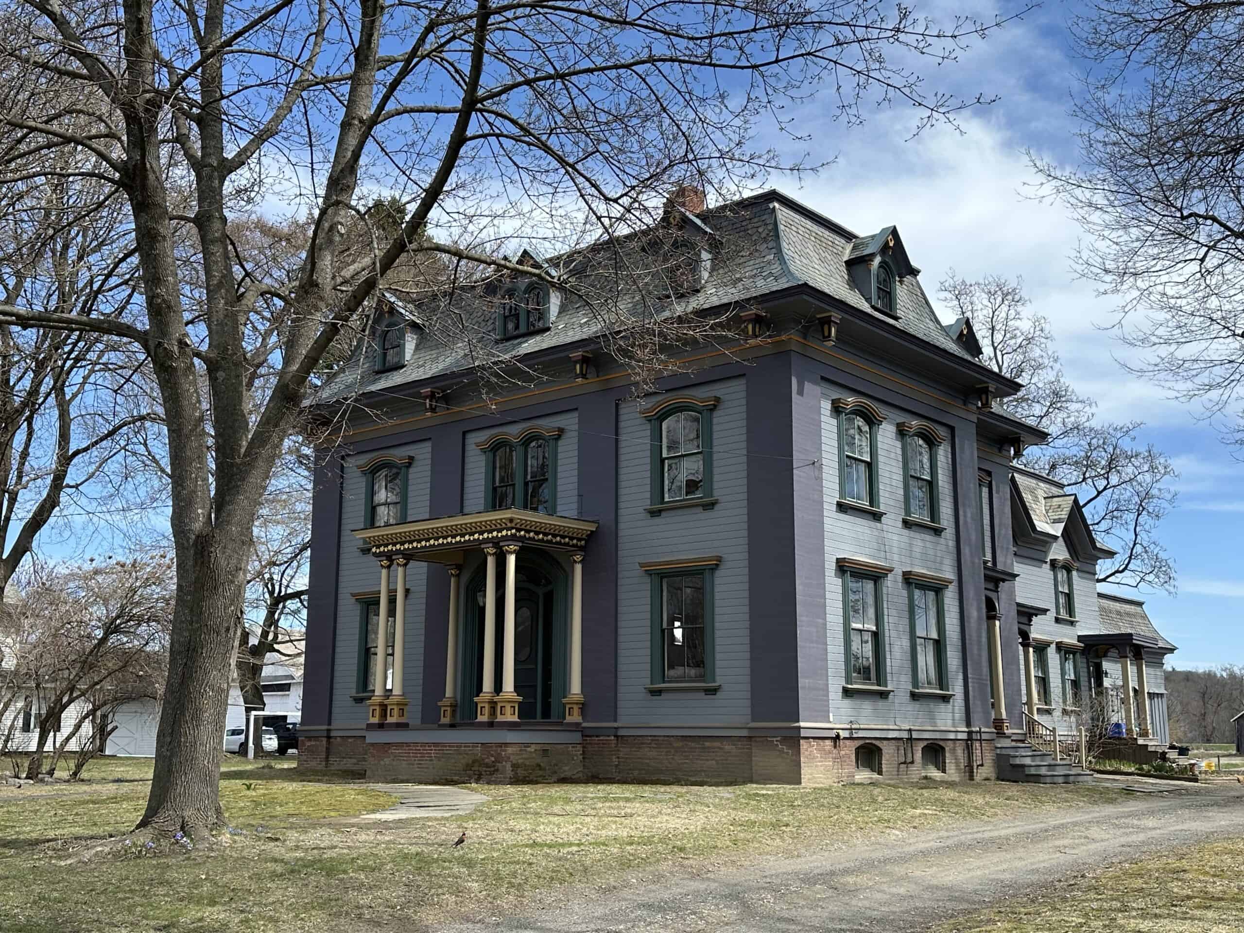 A large, three-story house featuring distinctive Second Empire and Italianate architectural elements. The structure is painted in a deep slate purple hue, contrasted by lighter grayish-blue panels on its facade and a brick foundation at the base. A prominent mansard roof, shingled in dark slate gray, crowns the building and is punctuated by multiple arched dormer windows with decorative trim. The windows across the upper floors are framed by ornate, carved wooden hoods painted in a contrasting brown or gold tone, matching the elaborate brackets supporting the overhanging cornices. On the left side of the facade, a grand, covered entrance portico stands supported by slender twin columns with decorative capitals. Bare, mature deciduous trees flank the left side of the property, casting thin shadows across the dormant grass lawn, while a gravel driveway curves along the right side toward the back of the house.