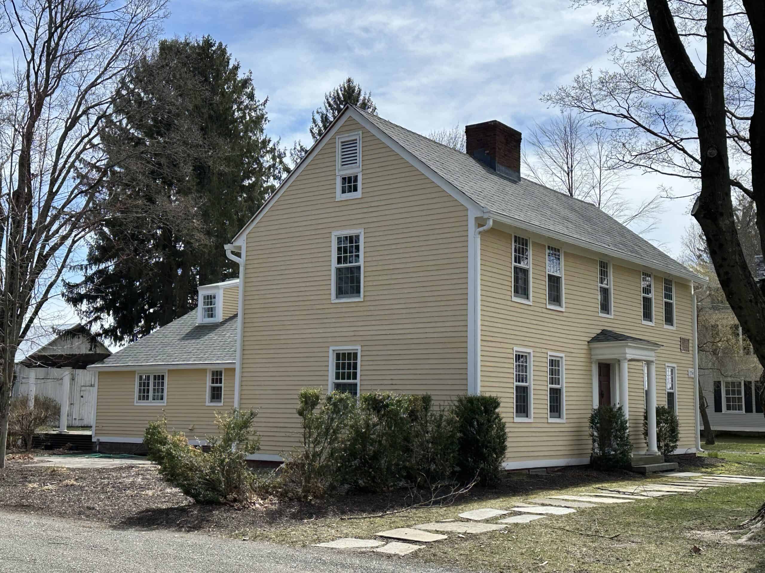 Light yellow, two-story colonial-style house with a steep gray-shingled gabled roof and white trim. The front entrance has a covered front portico supported by white columns. A large brick chimney rises from the center of the roof line. Attached to the left side of the main structure is a smaller, single-story addition with matching yellow siding, which extends toward the back of the property near a small detached white shed. The front yard contains trimmed green bushes, a stone slab walkway leading to the entrance, and mature, leafless trees framing the right side of the view.