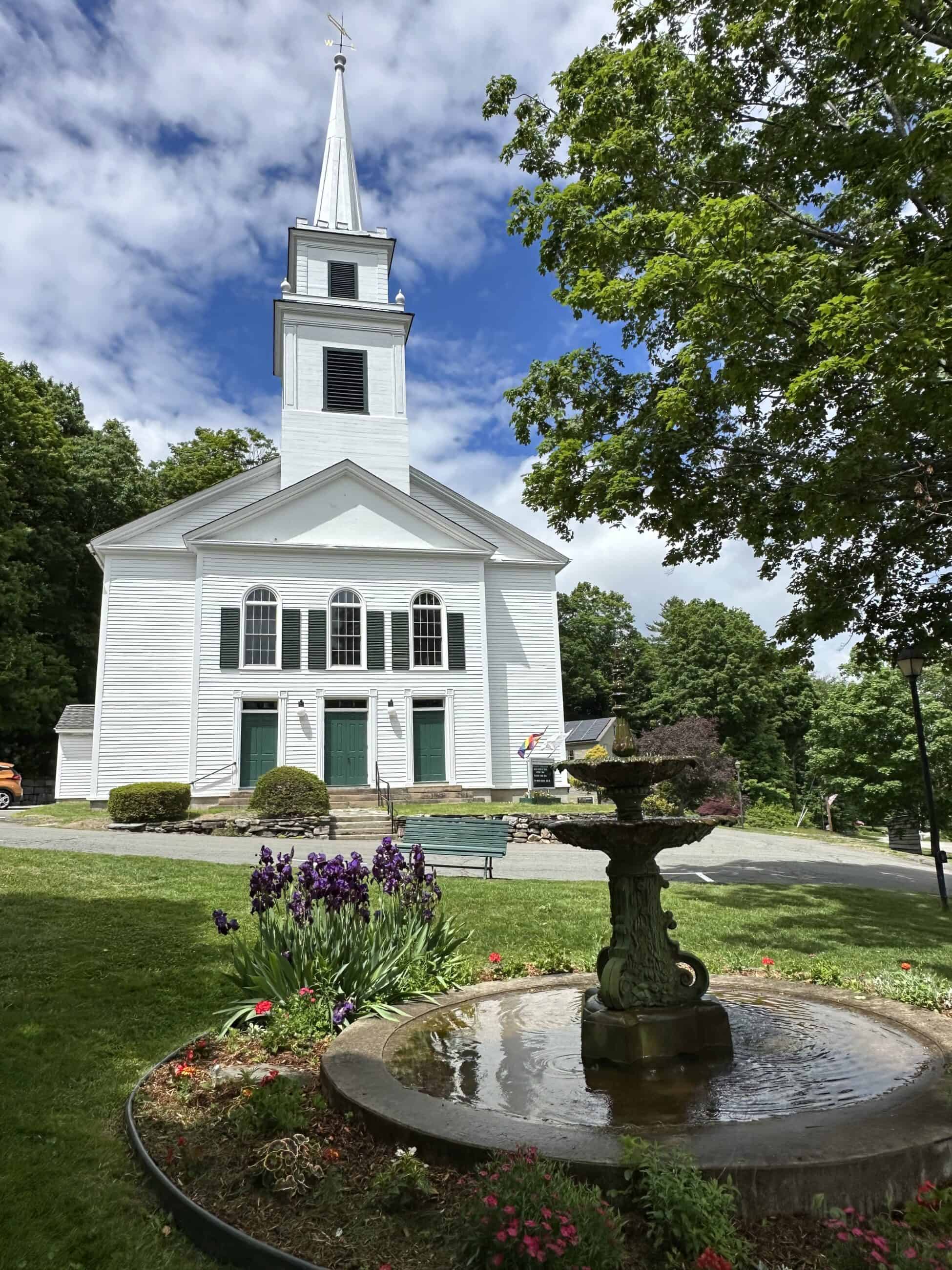 Modern photograph of the Westhampton Congregational Church. The church is white and in a simple meeting house style. Three green doors can be seen infront. In front of the house is a fountain surrounded by greenery and flowers.