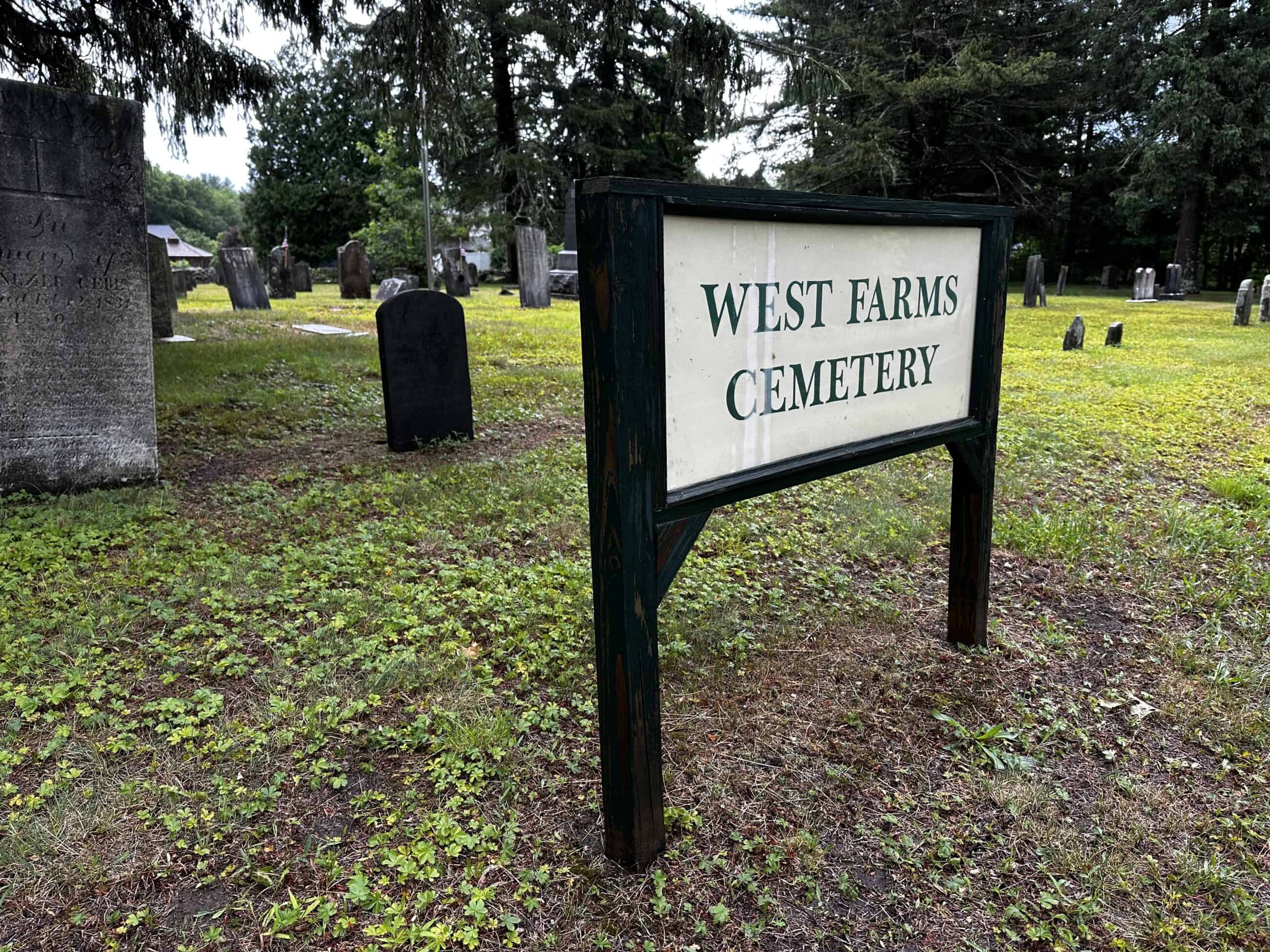 A wooden sign with a dark green or black painted frame stands in the foreground, displaying the text "WEST FARMS CEMETERY" in dark green capital letters on a light cream background. In the background are trees and old gravestones.