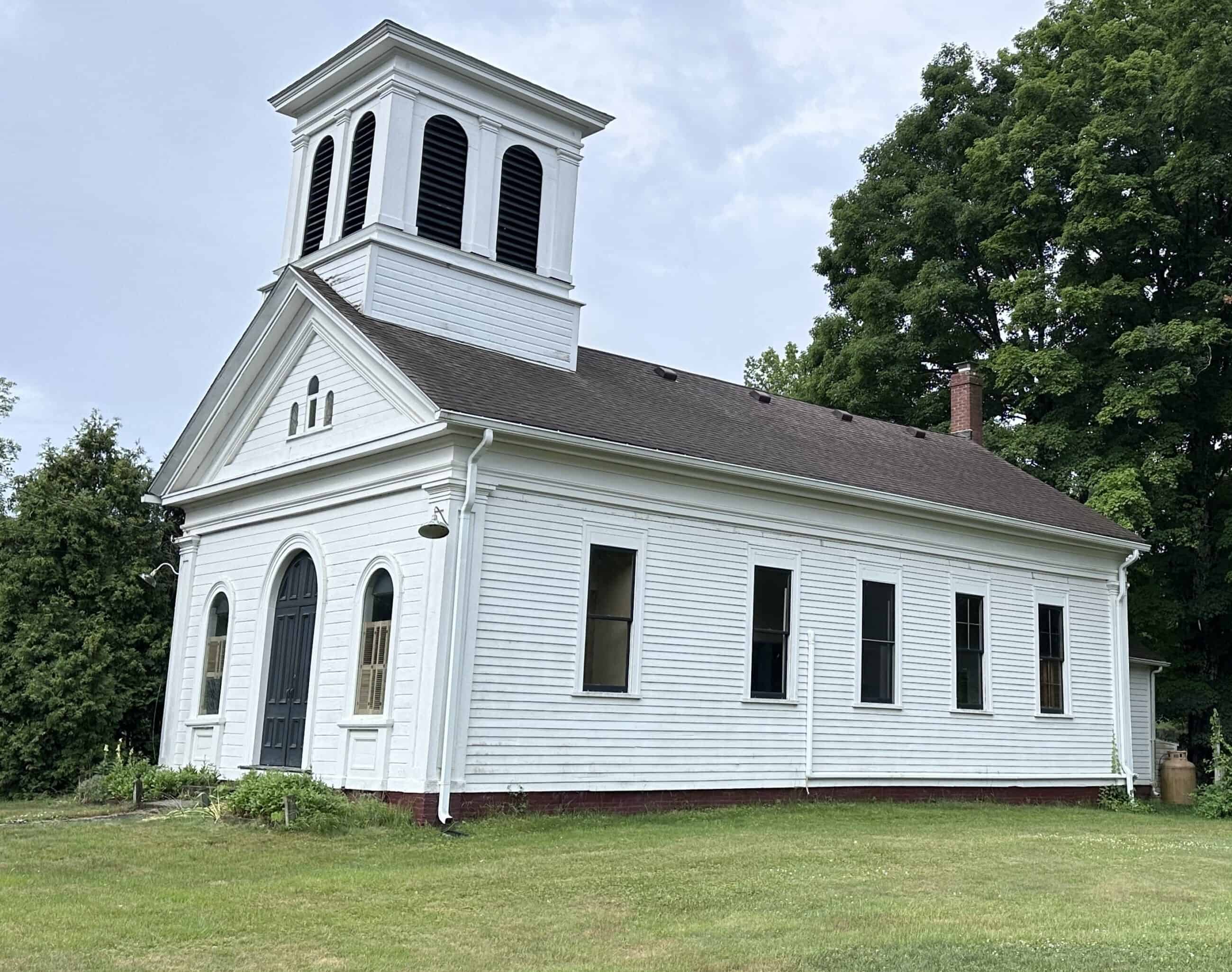 An exterior shot of a historic white wooden church building. The structure features a gabled roof and a prominent, square white bell tower or cupola rising from the front roofline, accented by black slatted louvers. The front facade includes a large, arched black double-door entrance flanked by tall, narrow arched windows. The long side wall displays a row of evenly spaced rectangular double-hung windows. Mature green trees stand prominently behind the church, and a grassy lawn surrounds the property.