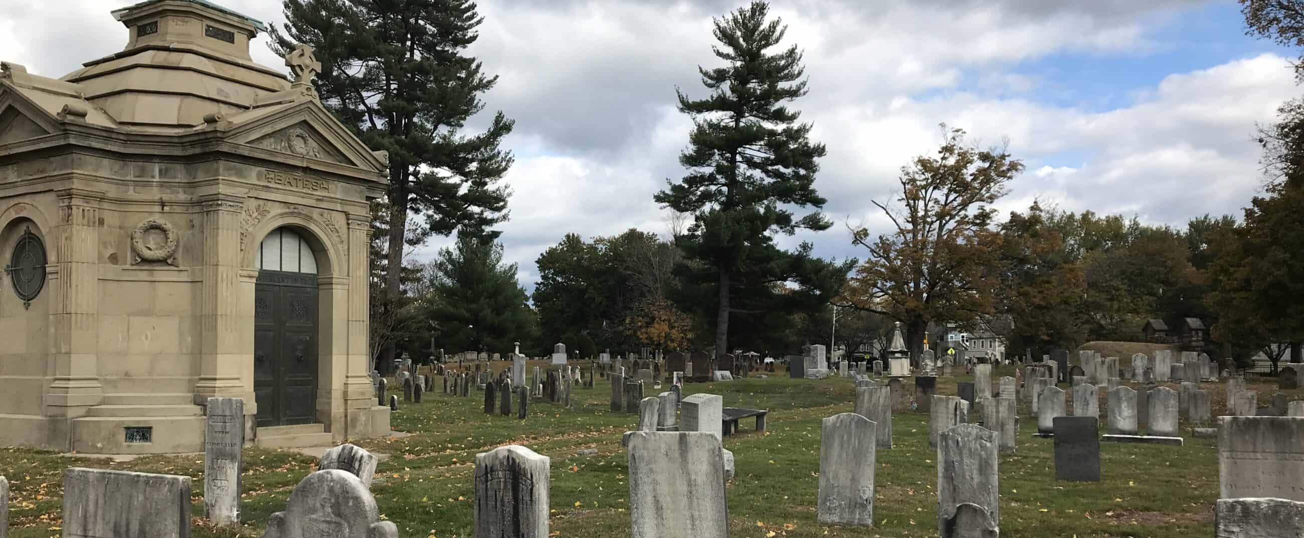 Photograph of the Bridge Street Cemetery. Old gravestones can be seen scattered around the grounds. Orange and yellow leaves are on the grass. An off-white, cream colored mausoleum is in view, engraved with the name "BATES."