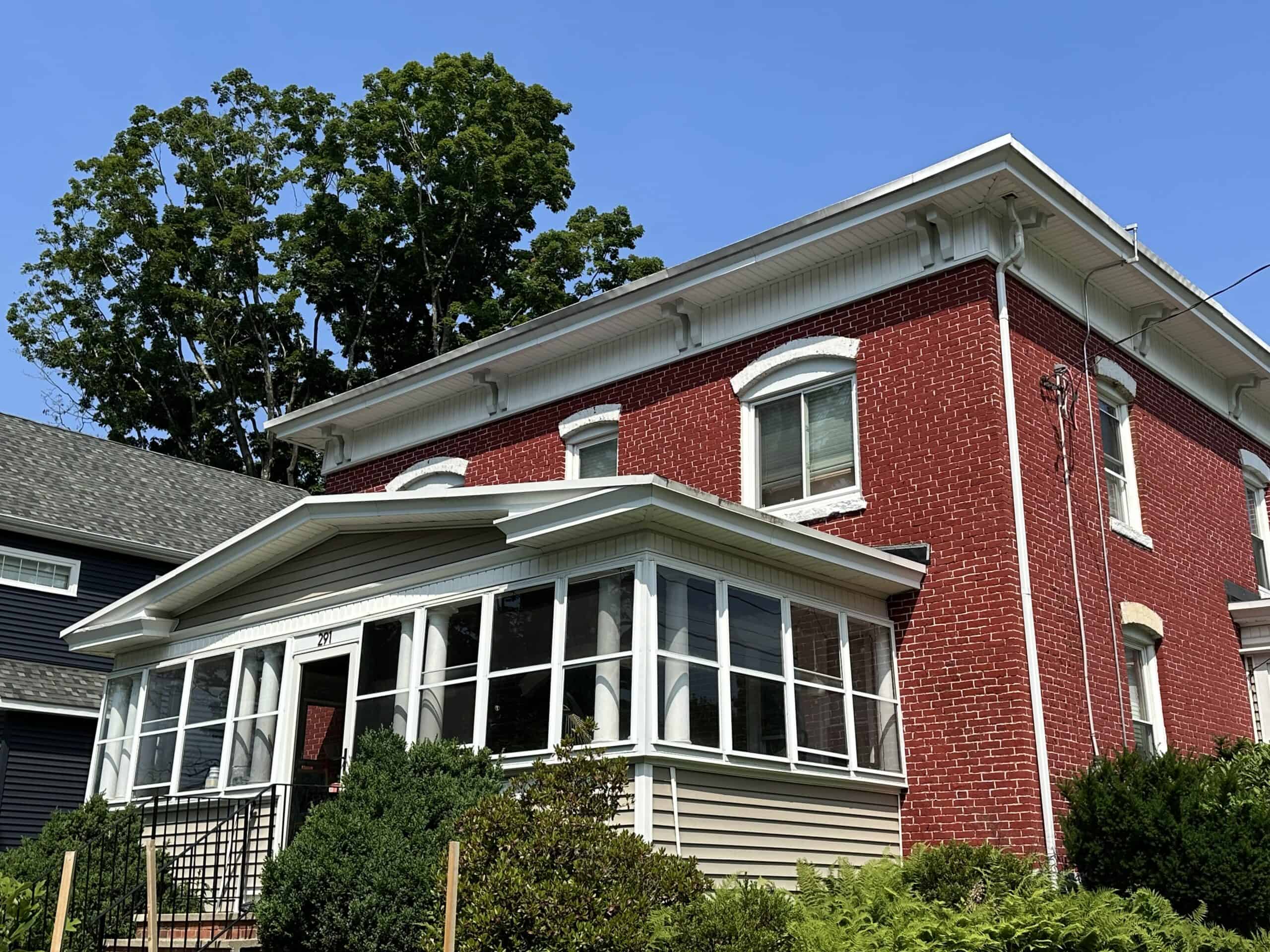 An image of a historic, two-story red brick building. The house features bright red brick walls with white trim along the roofline, which includes decorative white brackets beneath a wide overhang. On the second floor, multiple double-hung windows are visible, including one centered over the porch with a white curved decorative lintel above it, and two more on the right side of the house. The enclosed front porch, positioned on the lower left, has beige siding at its base, a peaked roof, and a row of large glass windows separated by white frames. A small set of stairs with a dark metal railing leads up to the porch entrance on the left. Green bushes and dense foliage grow in front of the porch and along the base of the house, partially obscuring the lower foundation. On the far left, a portion of an adjacent dark grey house with a shingled roof is visible. In the background, the lush green canopy of a tall tree rises above the roofline of the red brick house.