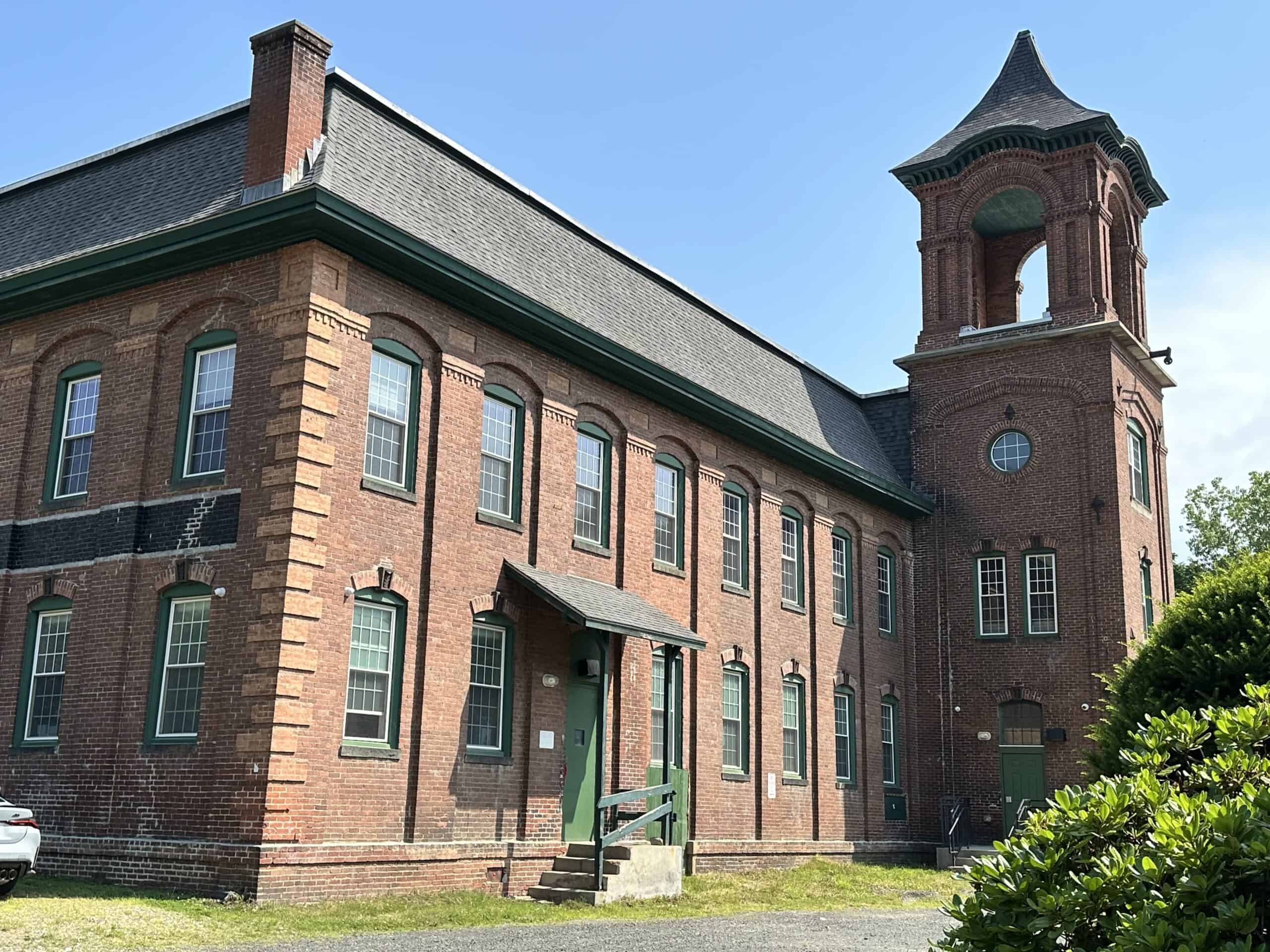 A three-quarter view of a historic, two-story red brick building. The structure features a dark grey roof with green trim. The left facade has tall, arched multi-pane windows with stone sills evenly spaced across both floors, along with vertical brick decorative detailing on the corner. A small awning with a dark roof and green support posts projects over a side entrance with a green door and concrete steps. To the right, a tall, attached square brick tower rises above the main building roofline. The tower has arched window openings at its top tier beneath a dark, flared bell-shaped roof, a small round window on the middle tier, and arched windows and a green door at ground level. A lush green bush frames the lower right foreground, while a small portion of a white car is visible on the far left.