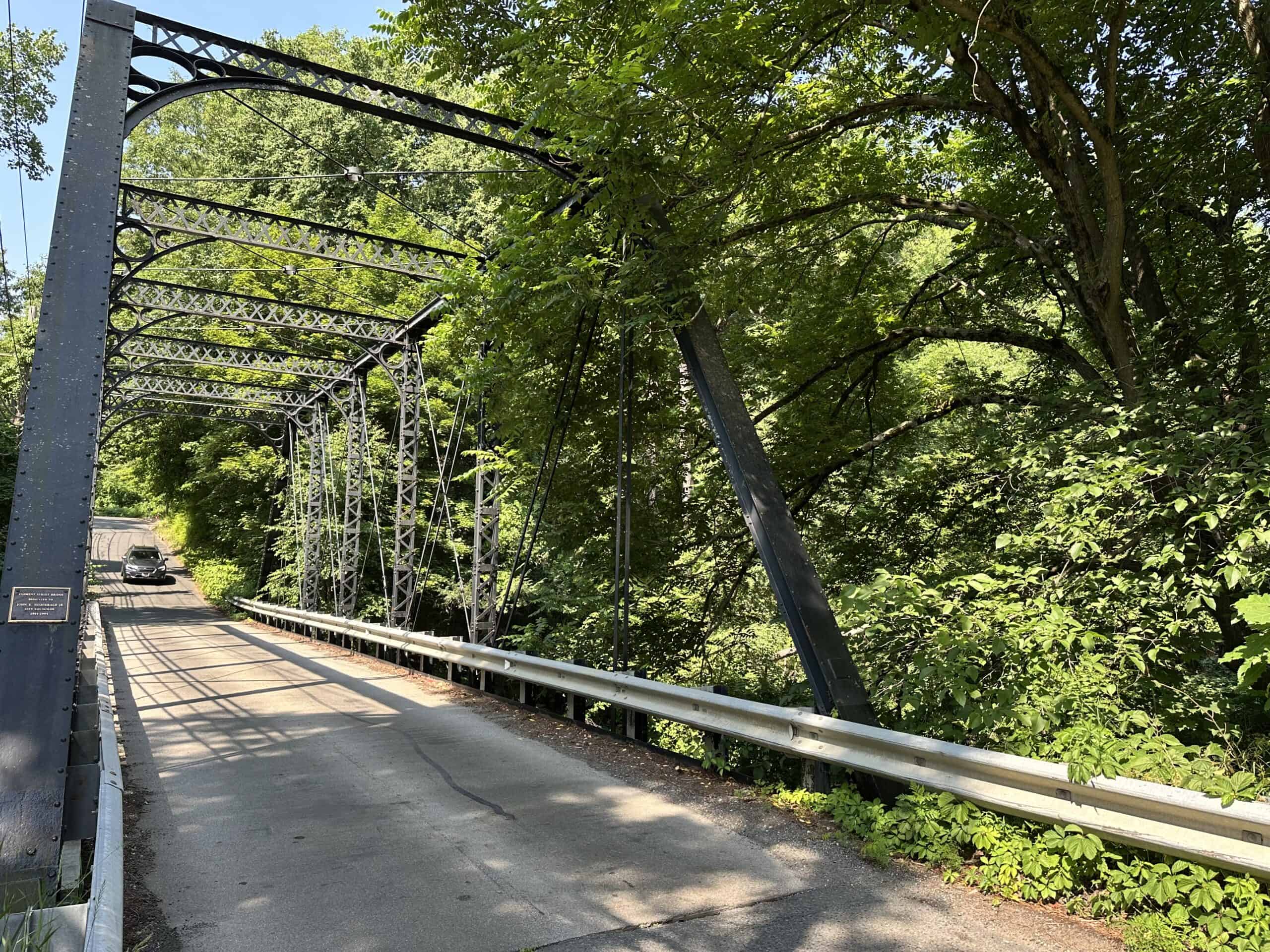 A photograph of a historic, single-lane bridge made of dark grey or black painted iron or steel. The bridge features an overhead, arched framework composed of latticed beams and horizontal cross-braces spanning the paved roadway. On the right side, dense, vibrant green foliage and mature trees grow closely against the structure, their branches leaning over the top of the bridge framework. A modern silver guardrail runs along the right edge of the paved surface, separating it from the steep drop-off into the unseen ravine or riverbank. In the background, the road curves gently upward and to the left, where a gray car is visible approaching the bridge from a distance. On the far left, a small, rectangular bronze plaque is affixed to the main vertical support beam of the bridge near the ground.