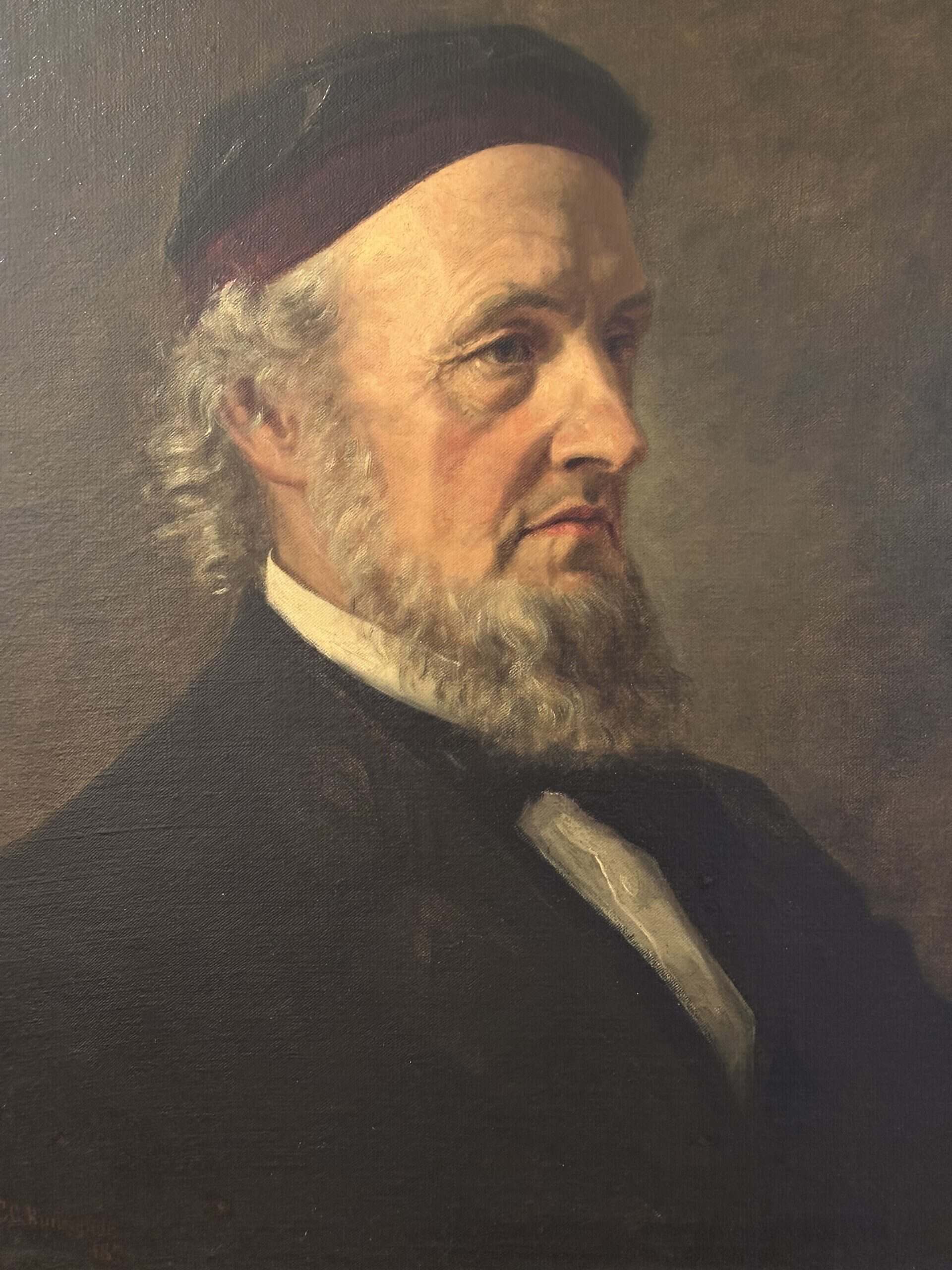 Portrait painting of a matured man with a white beard and white curly hair. He wears a black and purple cap on his head and a black and white suit and bow tie. The background is solid brown. The man looks past the audience off to the side and his facial expression is neutral.