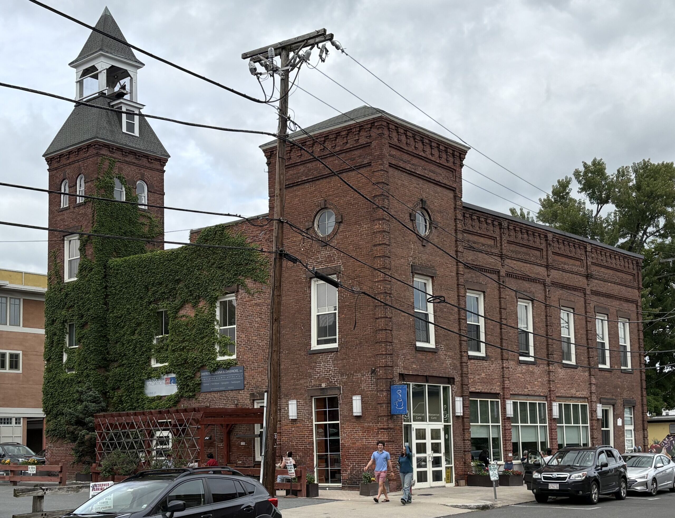 Photograph of a brick building with a bell tower. One side of the building is covered in ivy. Cars are parked on the side of the road and two people are walking by on the sidewalk.
