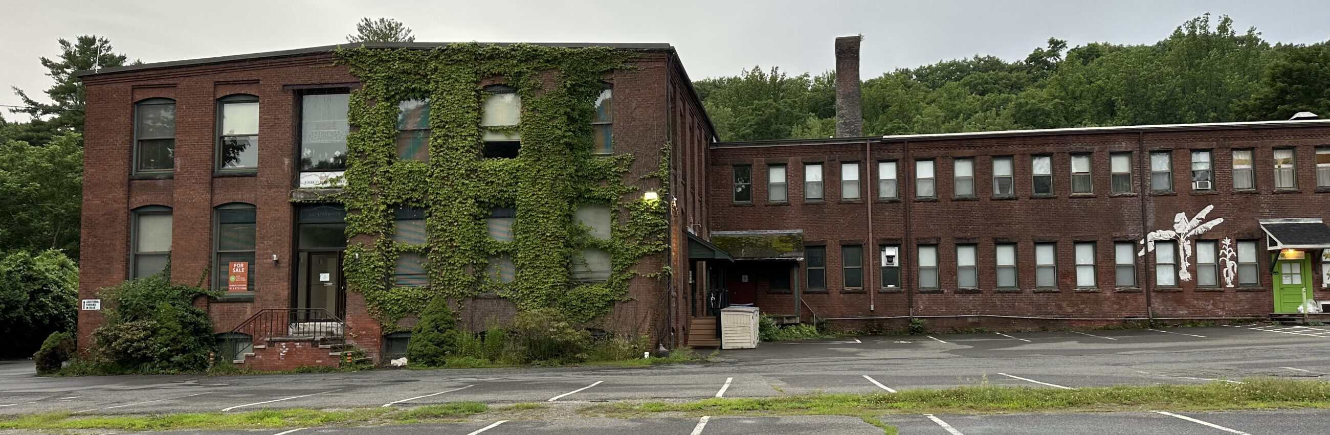 An image of a long, two-story industrial building made of dark red bricks under an overcast sky. The structure consists of two main sections: a taller block on the left with part of its facade densely covered in crawling green ivy, and an elongated right wing with rows of small, identical rectangular windows. A paved parking lot with faded white space markings stretches across the foreground, while a dense green forest covers the rising hillside immediately behind the property.