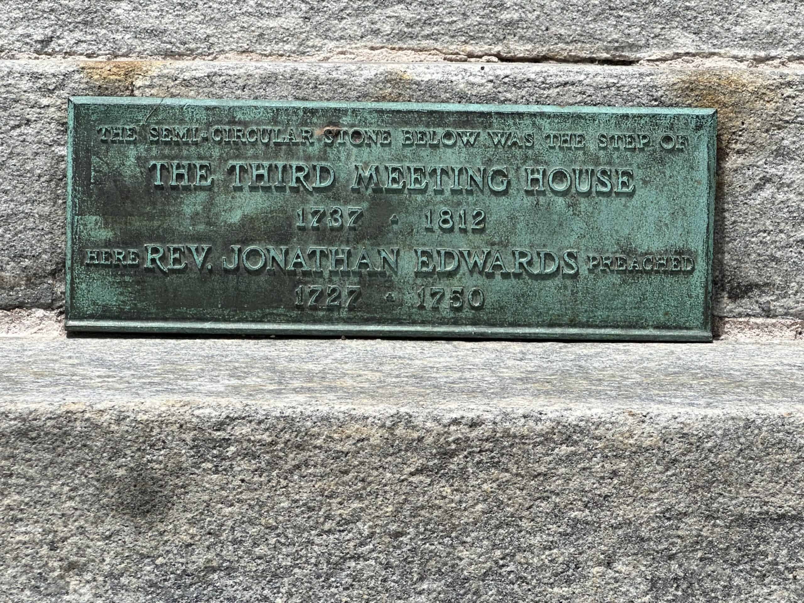 Photograph of the plaque representing the location of the old Third Meetinghouse. The plaque is embedded into a stone step. The plaque is a copperish green, and the engraving reads: "THE SEMI-CIRCULAR STONE BELOW WAS THE STEP OF THE THIRD MEETING HOUSE 1737 - 1812 HERE REV. JONATHAN EDWARDS PREACHED 1727 - 1750." 