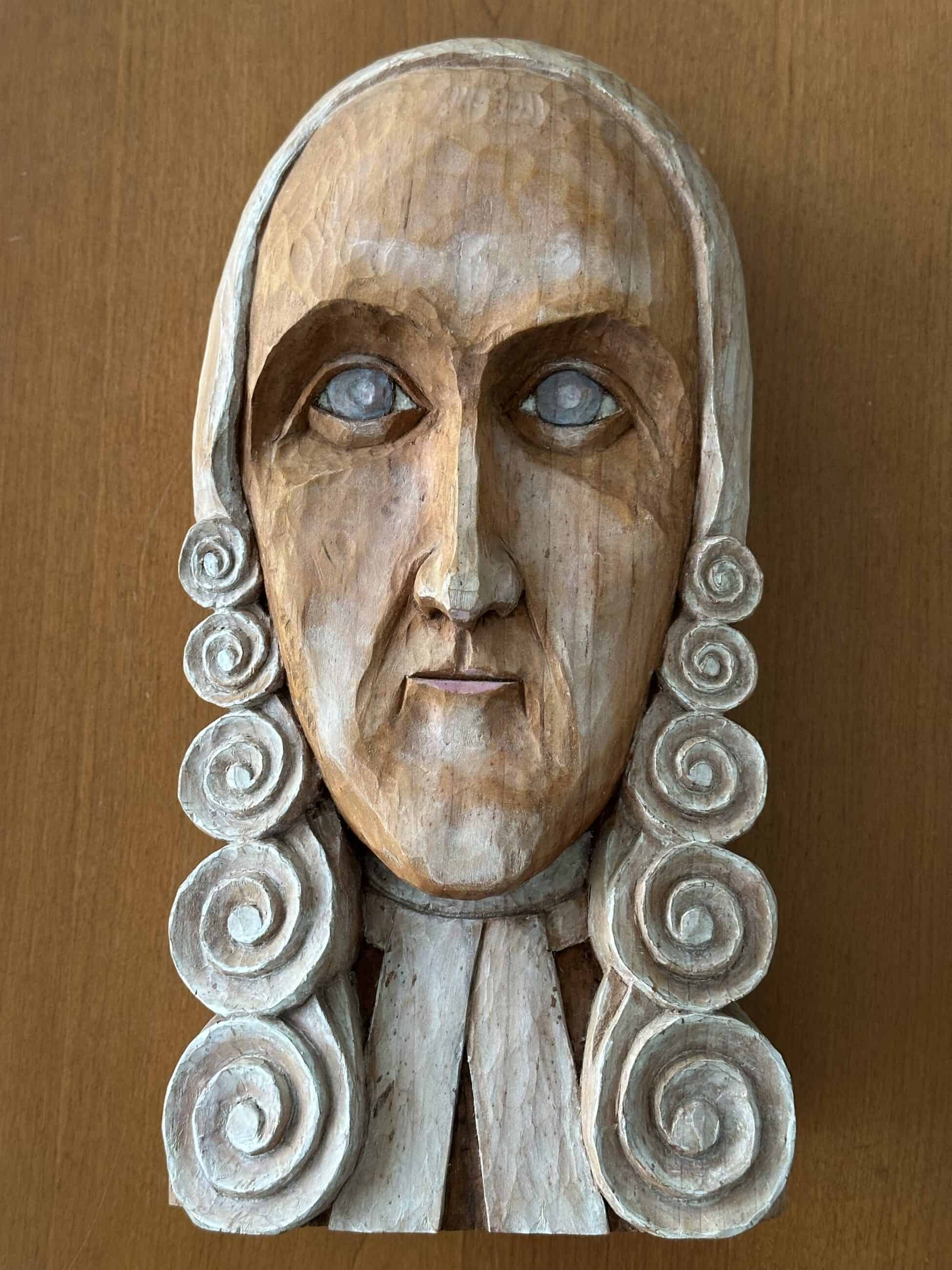 Wood sculpture of the face and tie of Jonathan Edwards. The sculpture is in a cartoonish style, with white curls styled like a wig framing his face. The paint is wearing, but Edwards' face is still a peachy color and his eyes appear to be faded blue. 