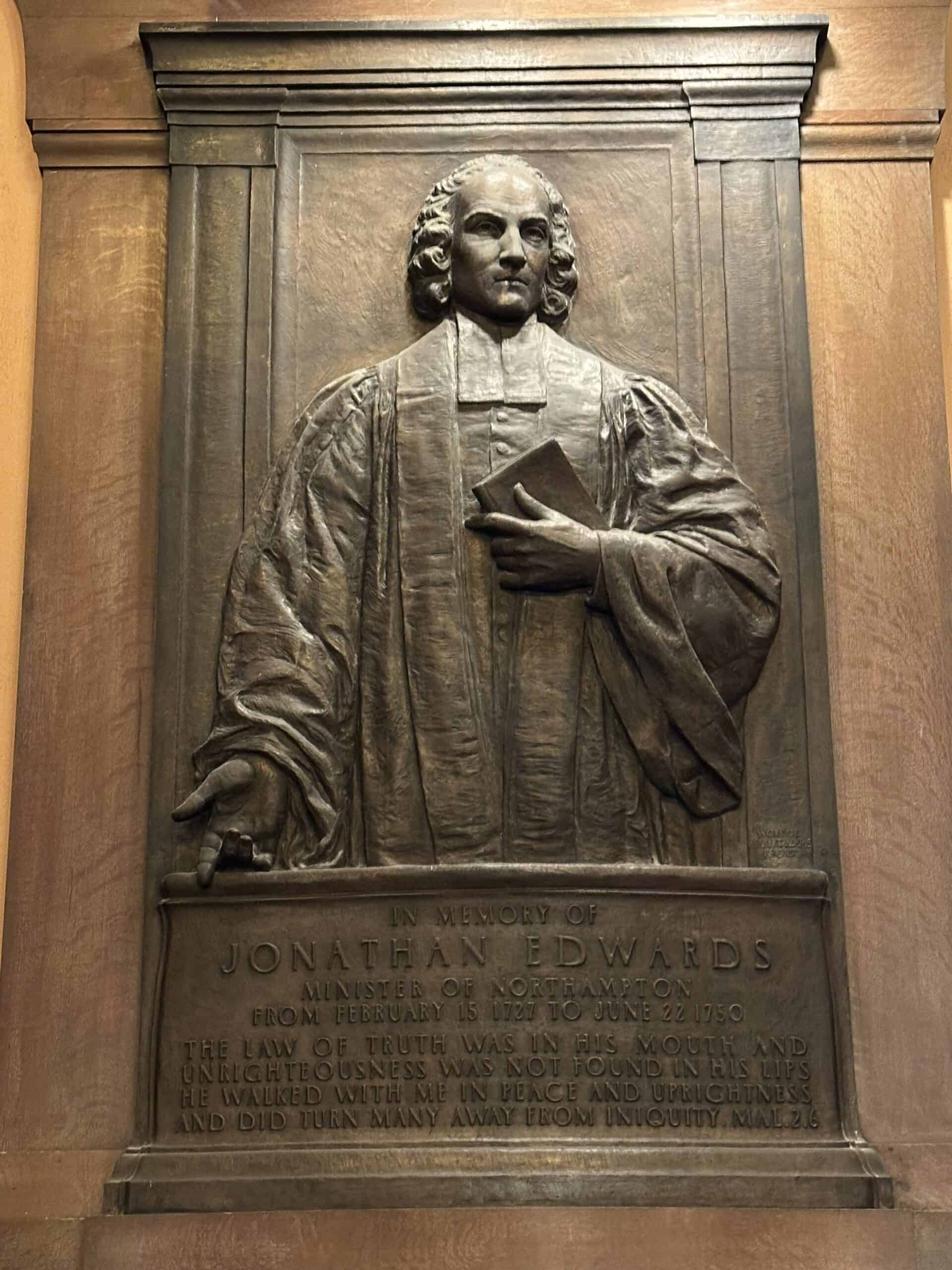 Photograph of the memorial tablet depicting Jonathan Edwards in the First Church of Northampton. Jonathan Edwards is drawn in a common, semi-realistic colonial portrait style, and he is holding a book to his chest with one hand. His other hand is pointing downward. He is cut off around the leg and below is a plaque styled like a pulpit. The engraving reads: "IN MEMORY OF JONATHAN EDWARDS MINISTER OF NORTHAMPTON FROM FEBRUARY 15 1727 TO JUNE 22 1750. THE LAW OF TRUTH WAS IN HIS MOUTH AND UNRIGHTEOUSNESS WAS NOT FOUND IN ITS LIPS. HE WALKED WITH ME IN PEACE AND UPRIGHTNESS AND DID TURN MANY AWAY FROM INIQUITY. MAL2:6."